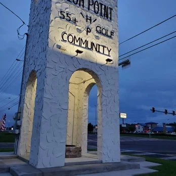 A white monument with a large archway and a rectangular top, displaying black text about a golf course community, with a parking lot and streetlights in the background during dusk.