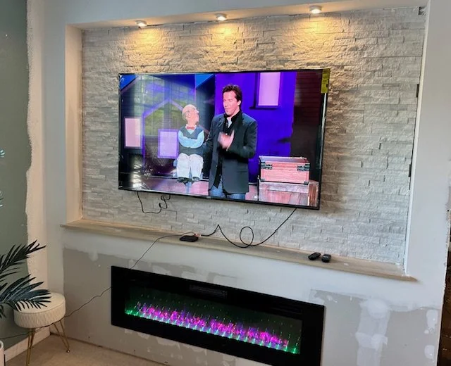 Flat-screen TV mounted on a textured white brick wall, displaying a show with two hosts, with a built-in electric fireplace below the TV.