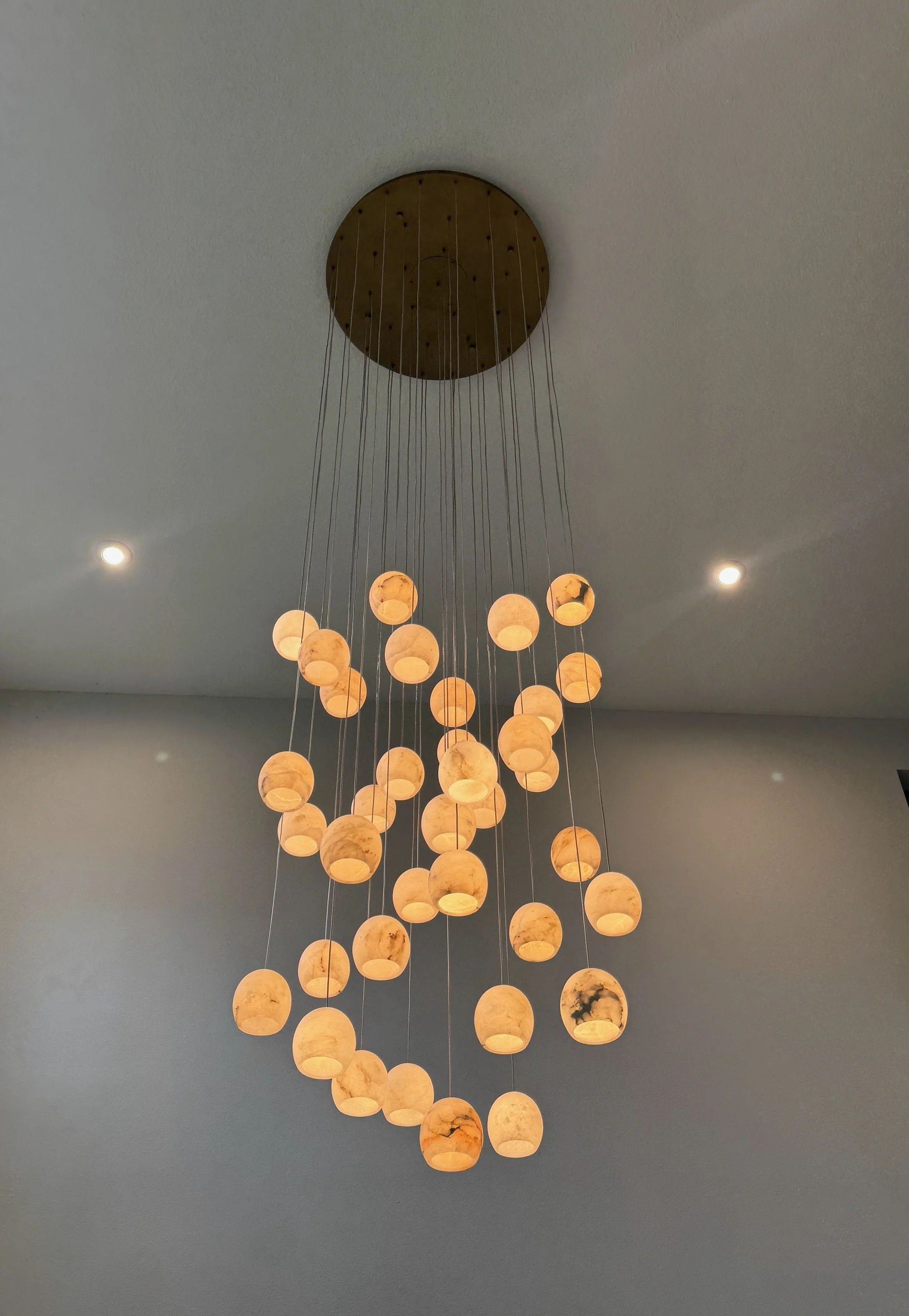 A modern cascading chandelier with multiple warm-glowing pendant lights suspended at varying lengths from a round ceiling mount. Each light features a soft, marble-like glass shade, creating an elegant waterfall effect and a warm, ambient glow agains