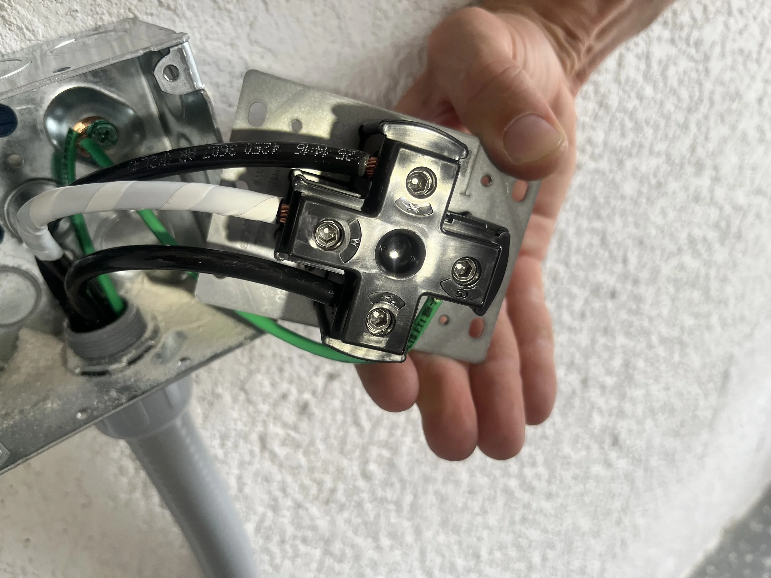 A person's hand holding electrical wiring connected to a metal electrical component mounted on a wall.