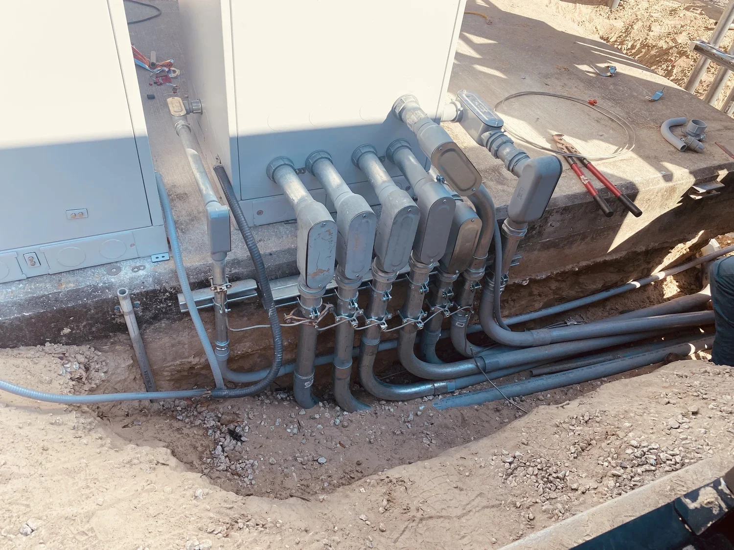 Construction site with electrical or plumbing pipes connected to a gray utility box on a concrete base, surrounded by dirt and construction tools.