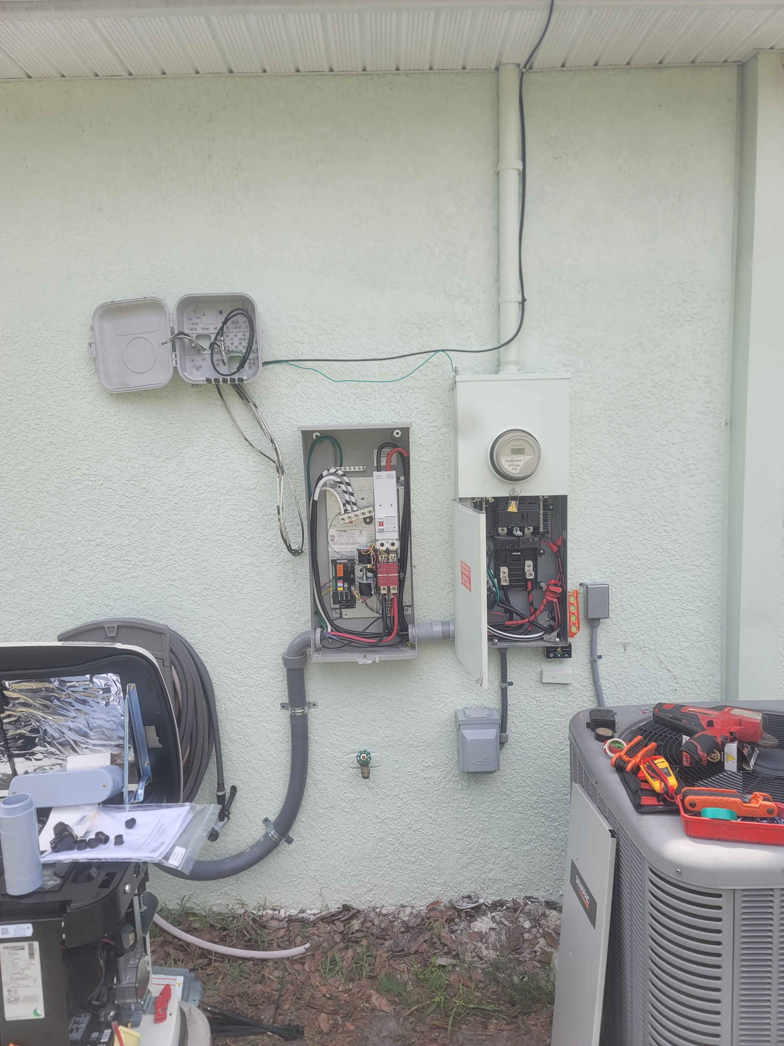 Exterior view of electrical and HVAC equipment, including open electrical panels, a power meter, and tools on a worktable, against a light-colored wall.