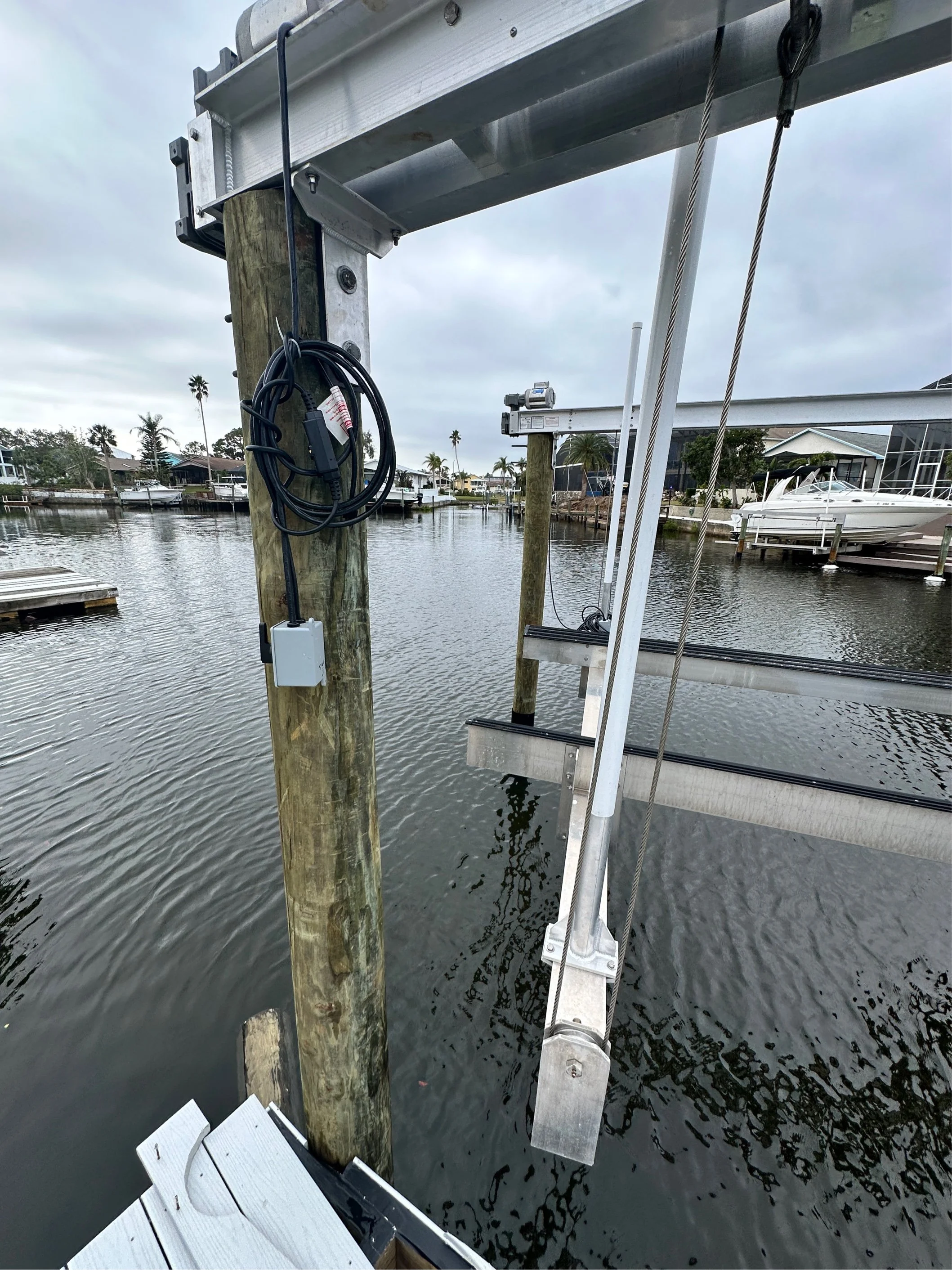 Close-up of a boat lift in a marina with metal framework, wooden posts, and electrical wiring; water with visible ripples below and boats in the background under an overcast sky.