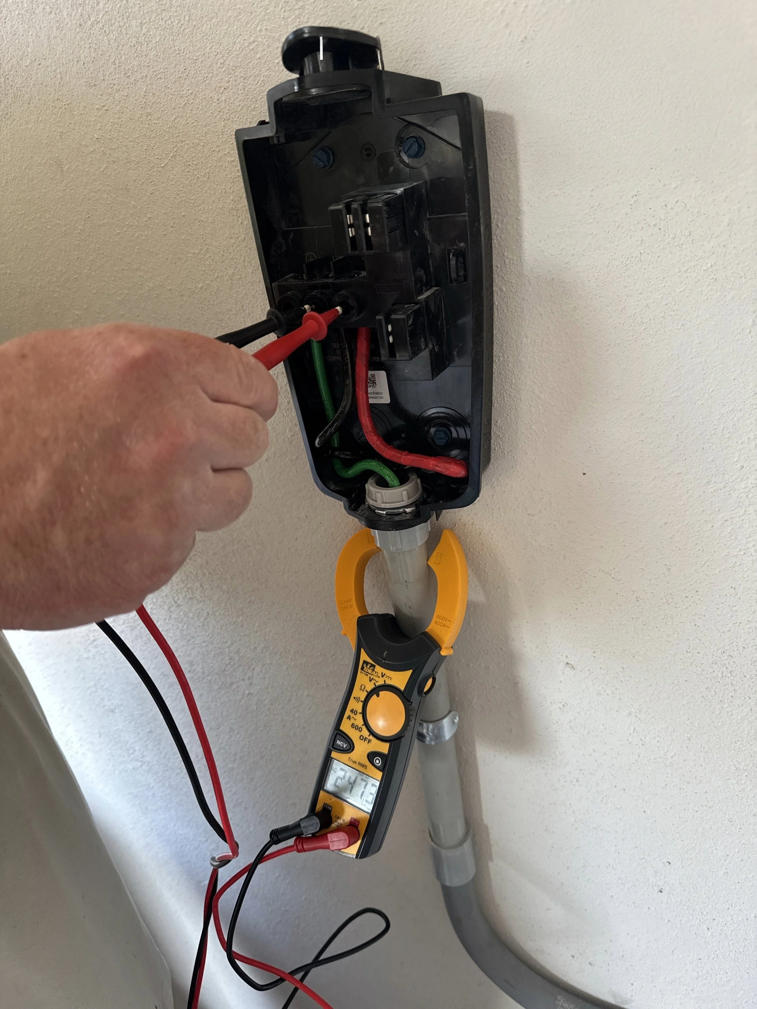 A person using a digital multimeter to test electrical wires inside an open electric vehicle box mounted on a wall.
