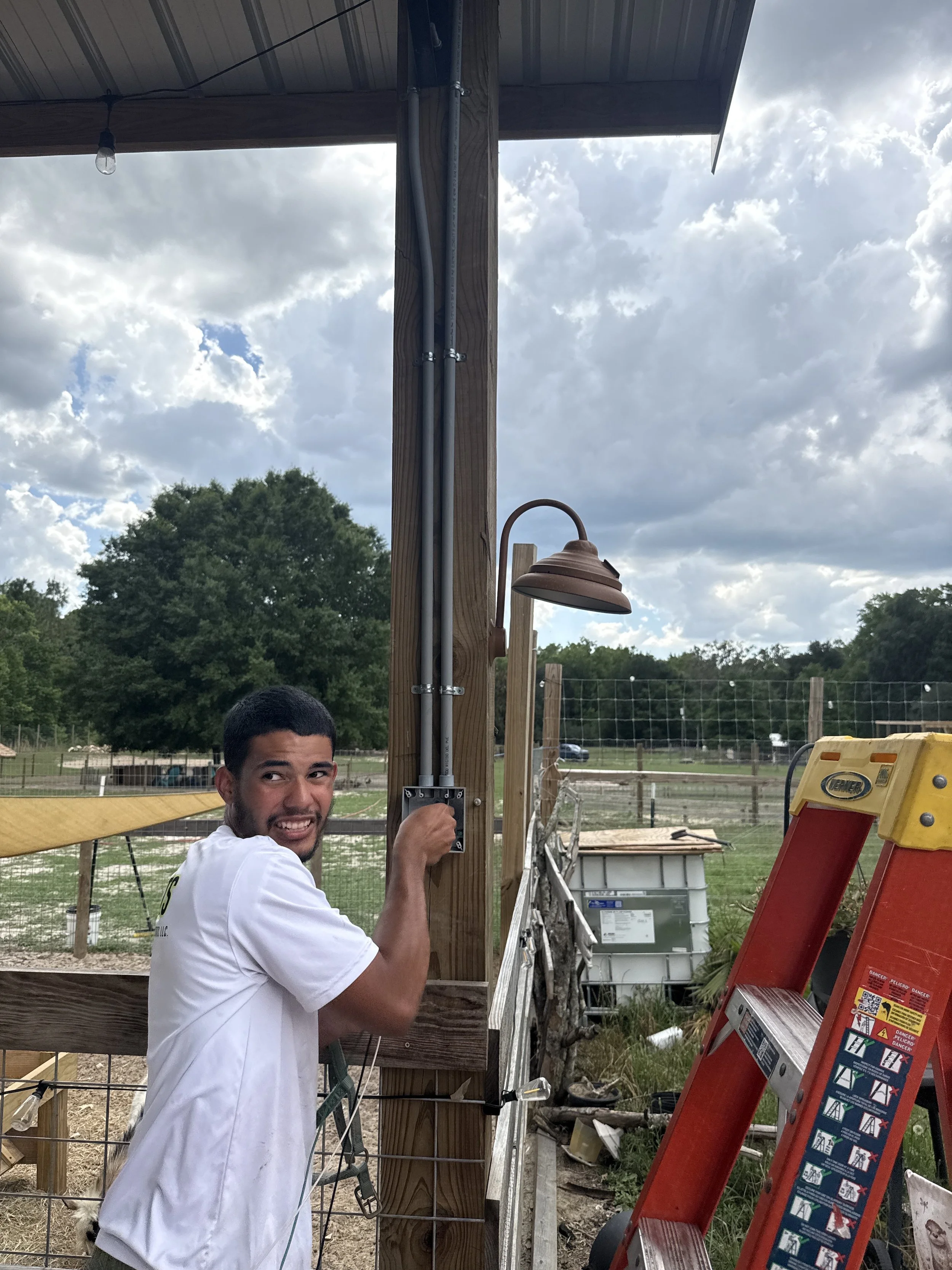 A young man wearing a white shirt is working on installing electrical wiring on a wooden fence post outdoors. There is a ladder nearby, a red extension ladder with warning labels, and a wooden structure with a small copper outdoor light fixture. The background shows trees, a field, and a partly cloudy sky.