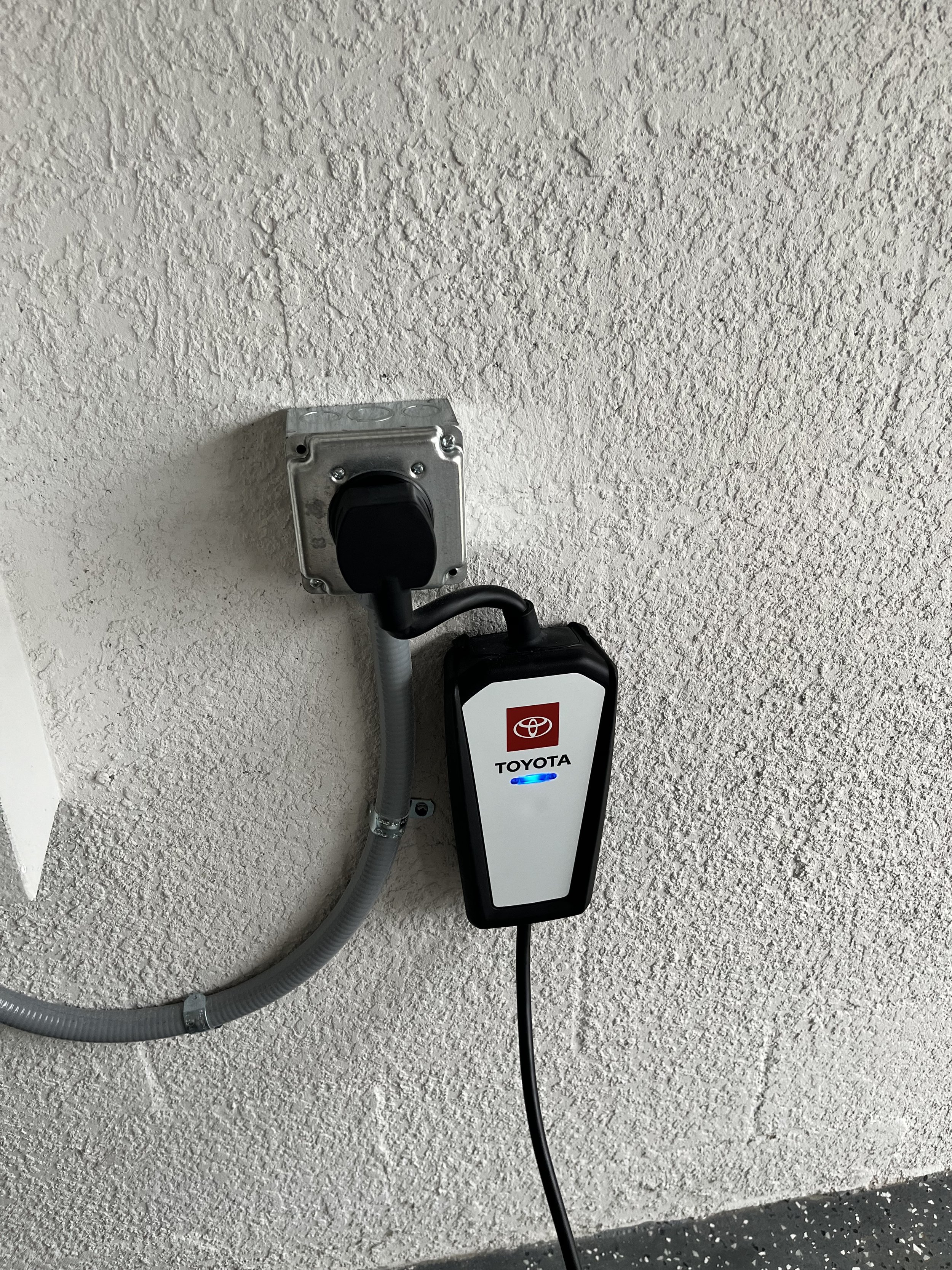 Electric vehicle charging station attached to a textured wall, with a black cable plugged into it, labeled with Toyota logo.
