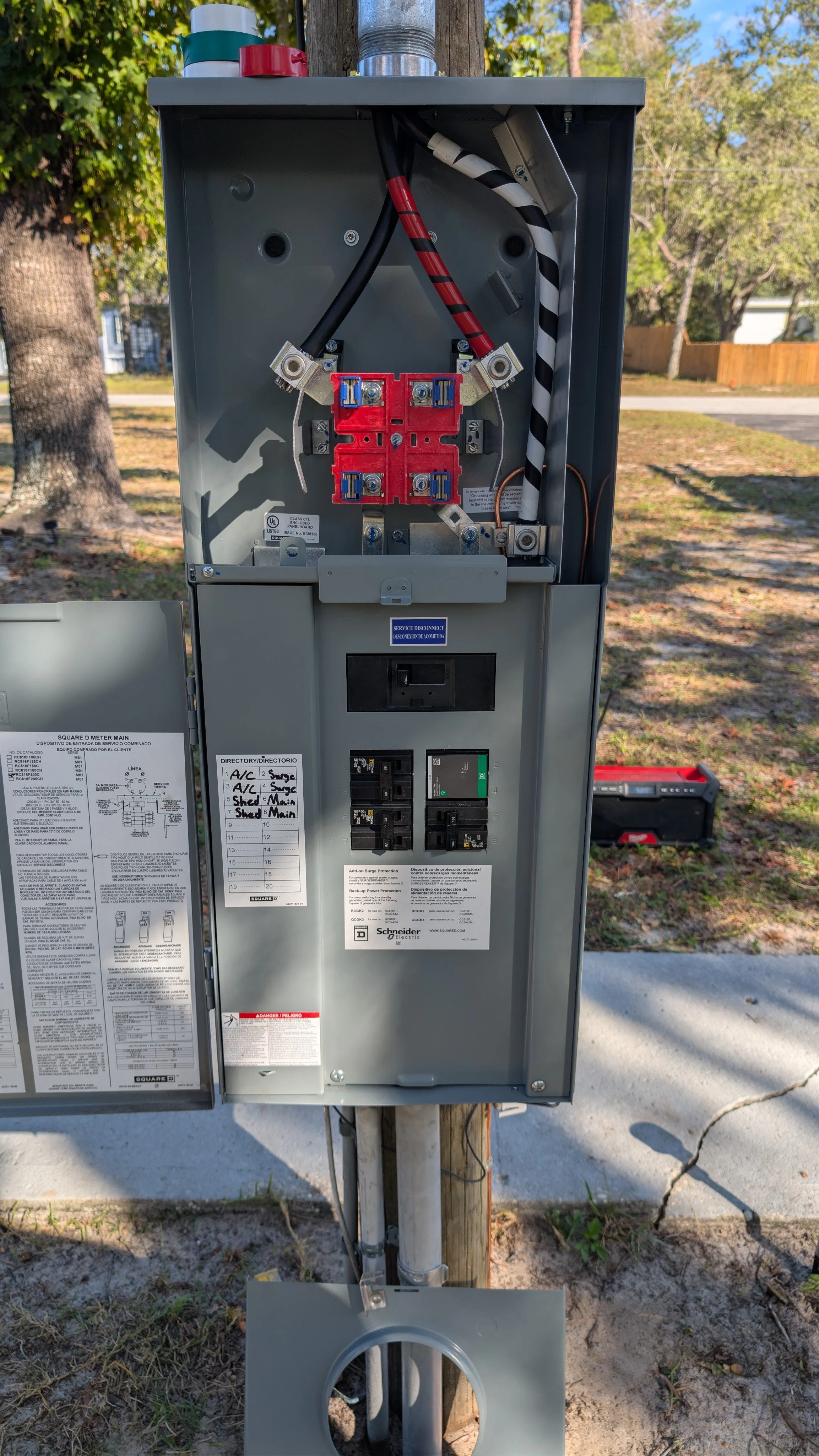 Electrical utility meter and breaker box mounted on a metal pole, with wiring and labels, outdoors on a sidewalk with trees and houses in the background.