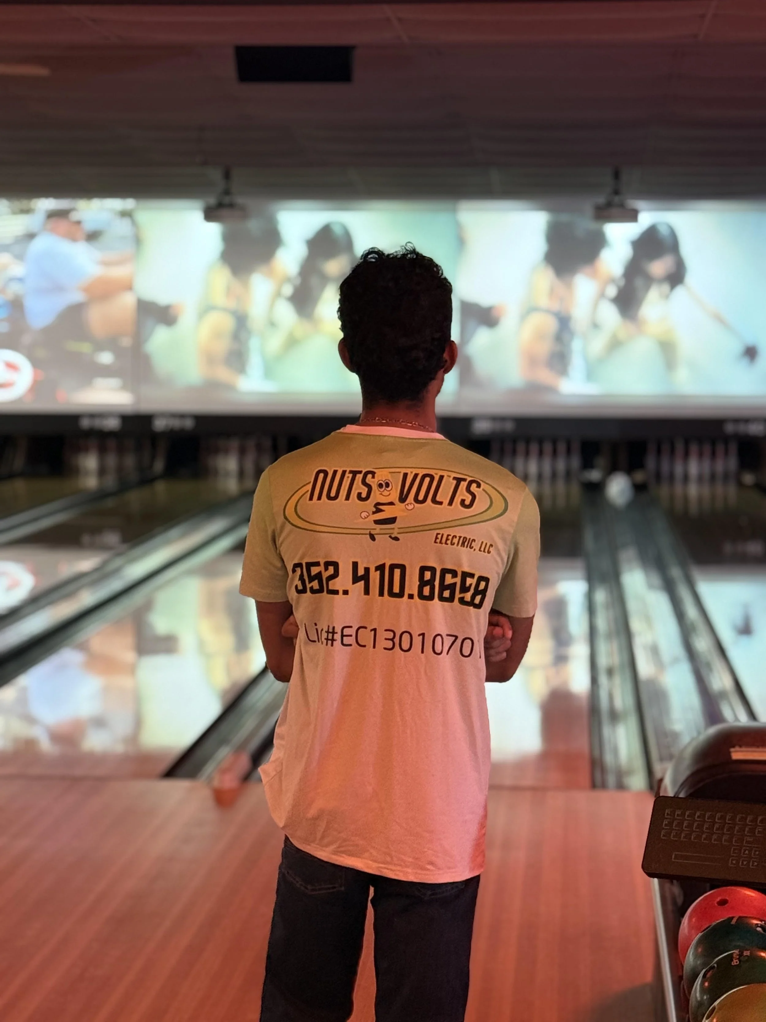 A person with curly hair wearing a yellow t-shirt with business contact information, standing in a bowling alley, facing the bowling lanes and screens.