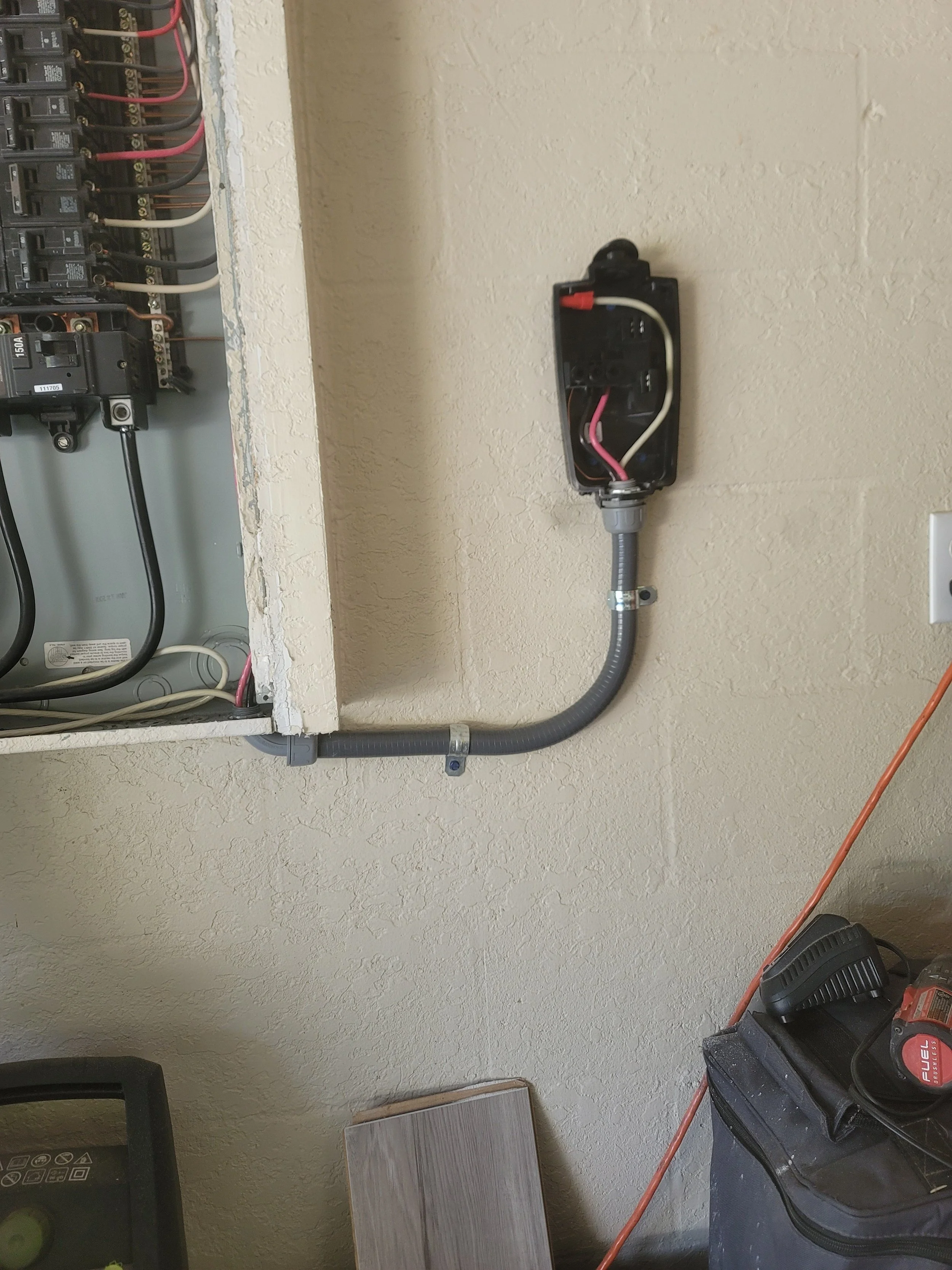Electrical panel with wiring and a disconnected outlet box with wiring, a conduit pipe connecting the outlet box to the panel, and tools on a nearby surface.