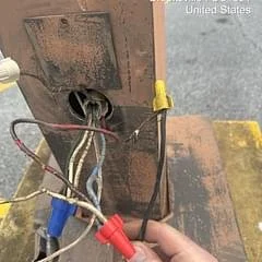 A person's hand holding a bundle of multicolored wires and connectors near an open electrical box on a wooden surface.