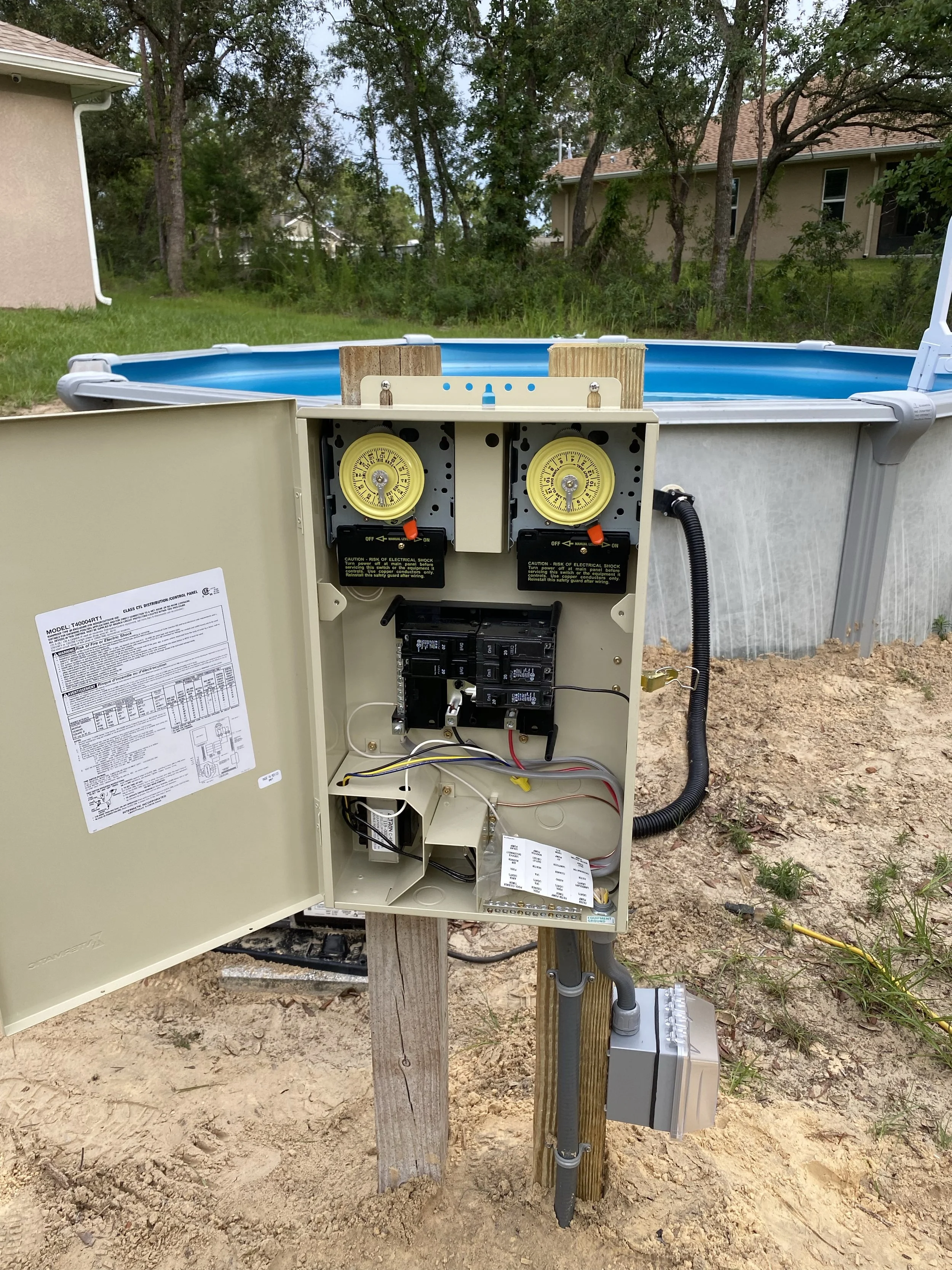 An open electrical box with timers and wiring, mounted on a wooden post next to an above ground pool in a backyard.