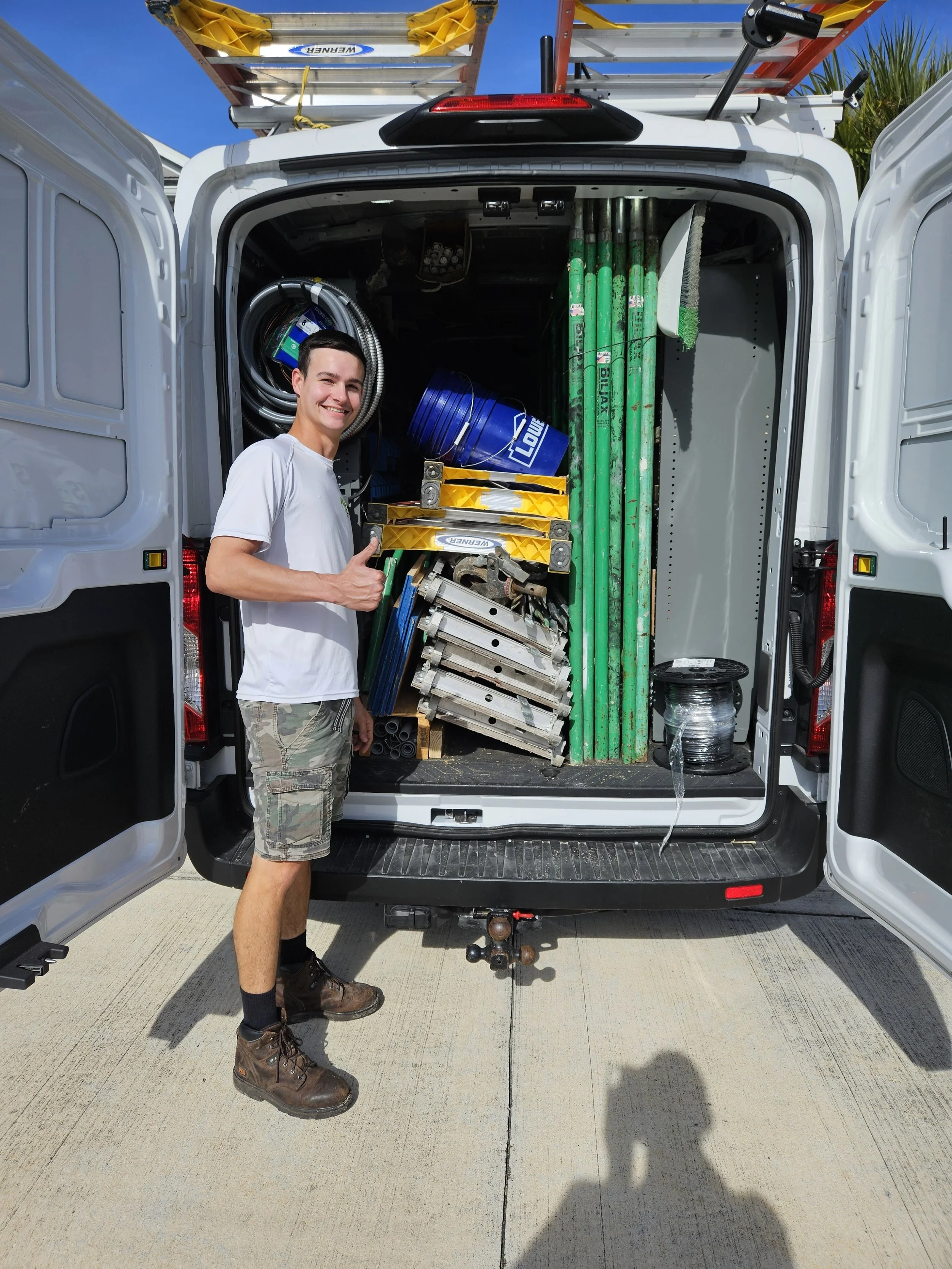 A smiling man in white t-shirt and camouflage shorts giving a thumbs up next to an open van loaded with maintenance equipment like pipes, ladders, and wiring.