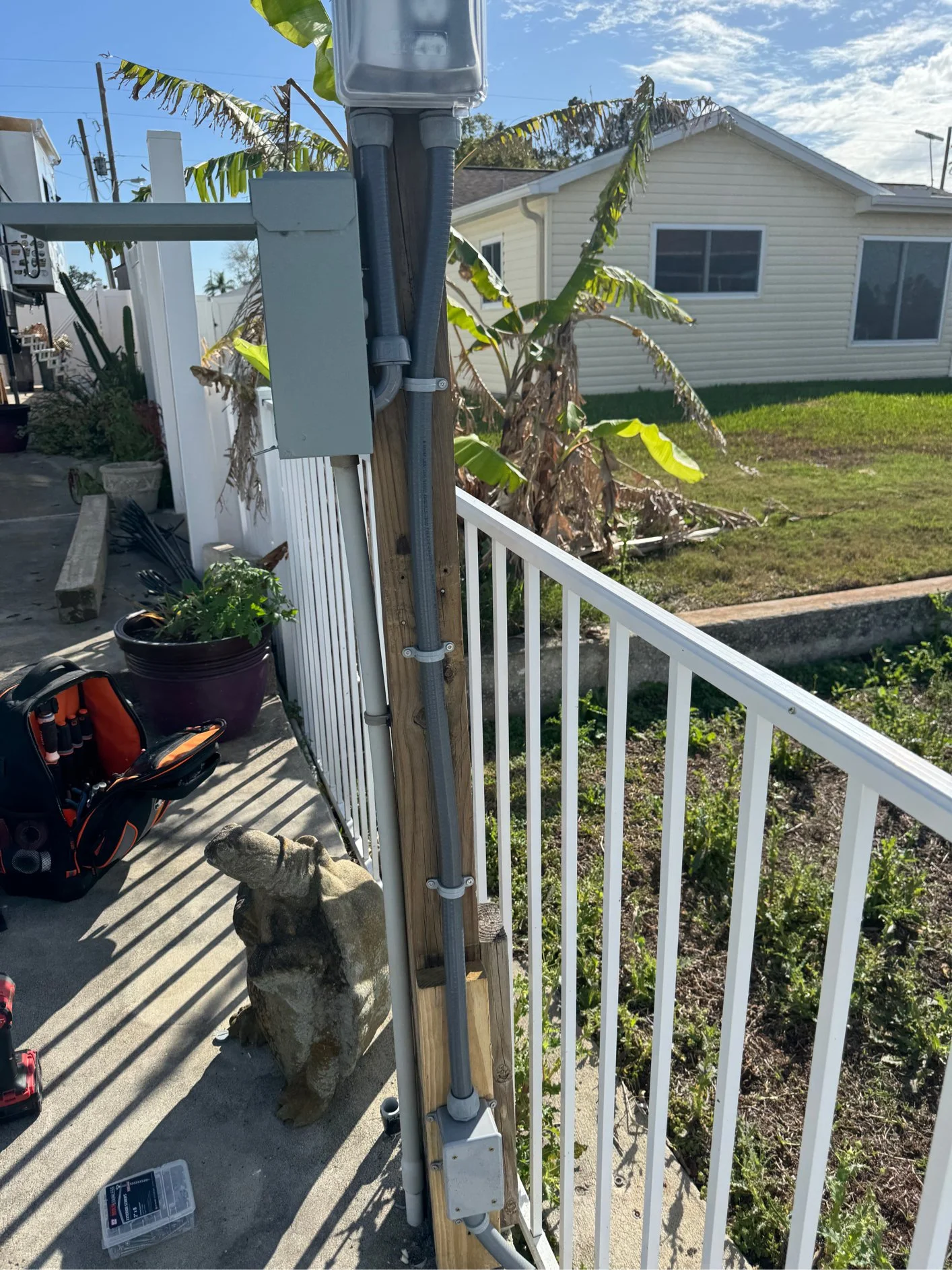 Close-up of an electrical conduit and junction box attached to a wooden post on a porch, with a dog figurine, garden tools, and a toolbox nearby, with a house, plants, and banana trees in the background.