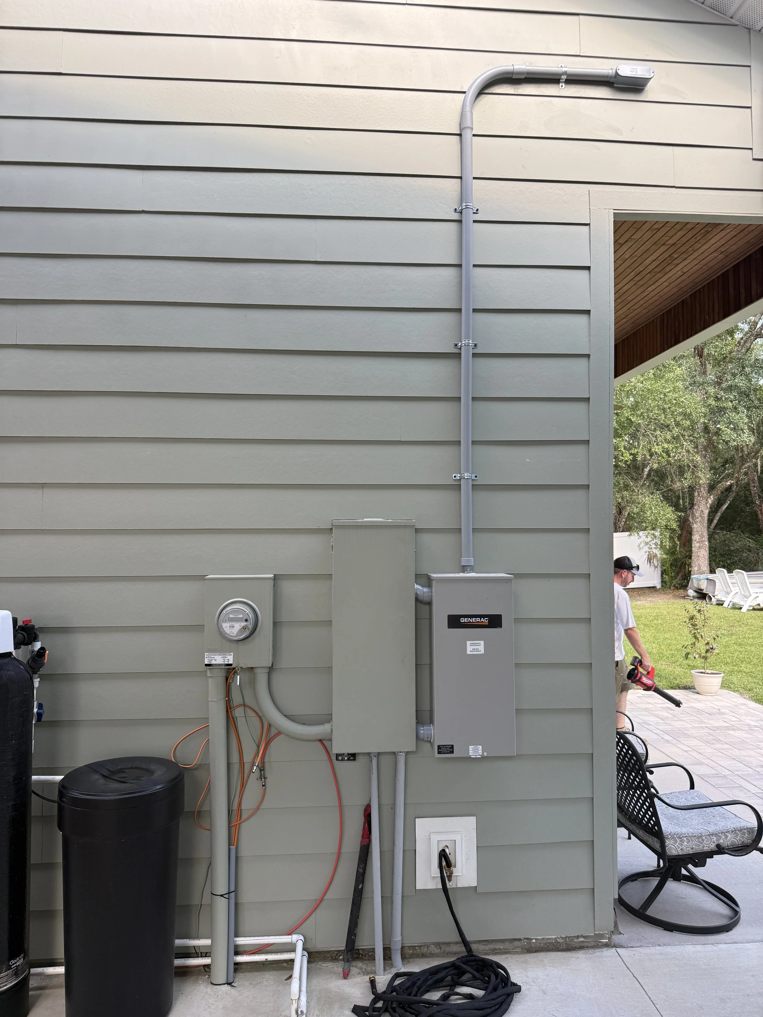Outside view of a house wall with electrical and plumbing installations, including a gray electrical panel, circuit meter, and conduits. There is a person walking on the patio wearing sunglasses and a white shirt, carrying a leaf blower.