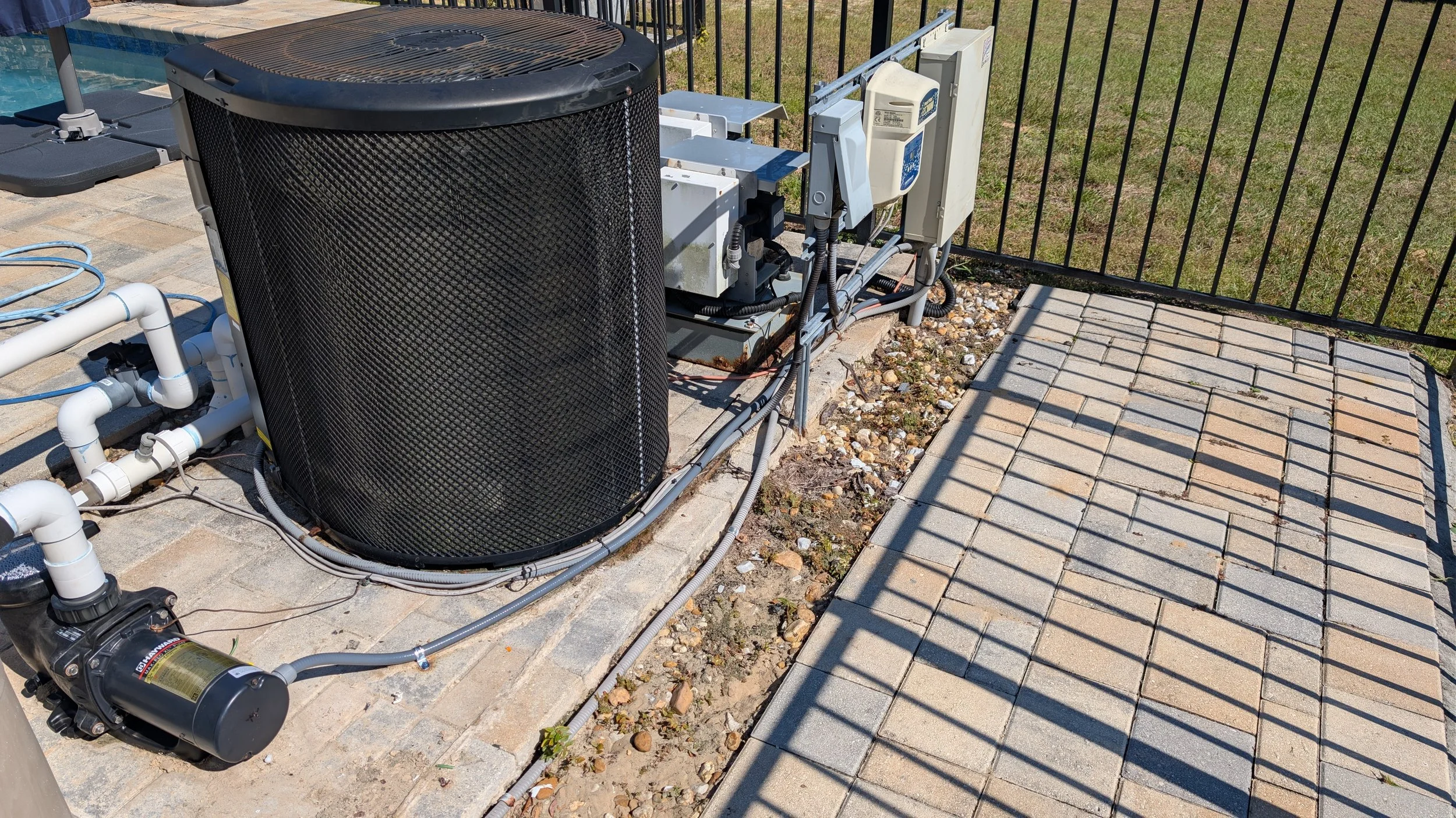 Outdoor air conditioning unit and pump on a concrete slab near a black metal fence with brick-paved walkway, grass, and pool in the background.