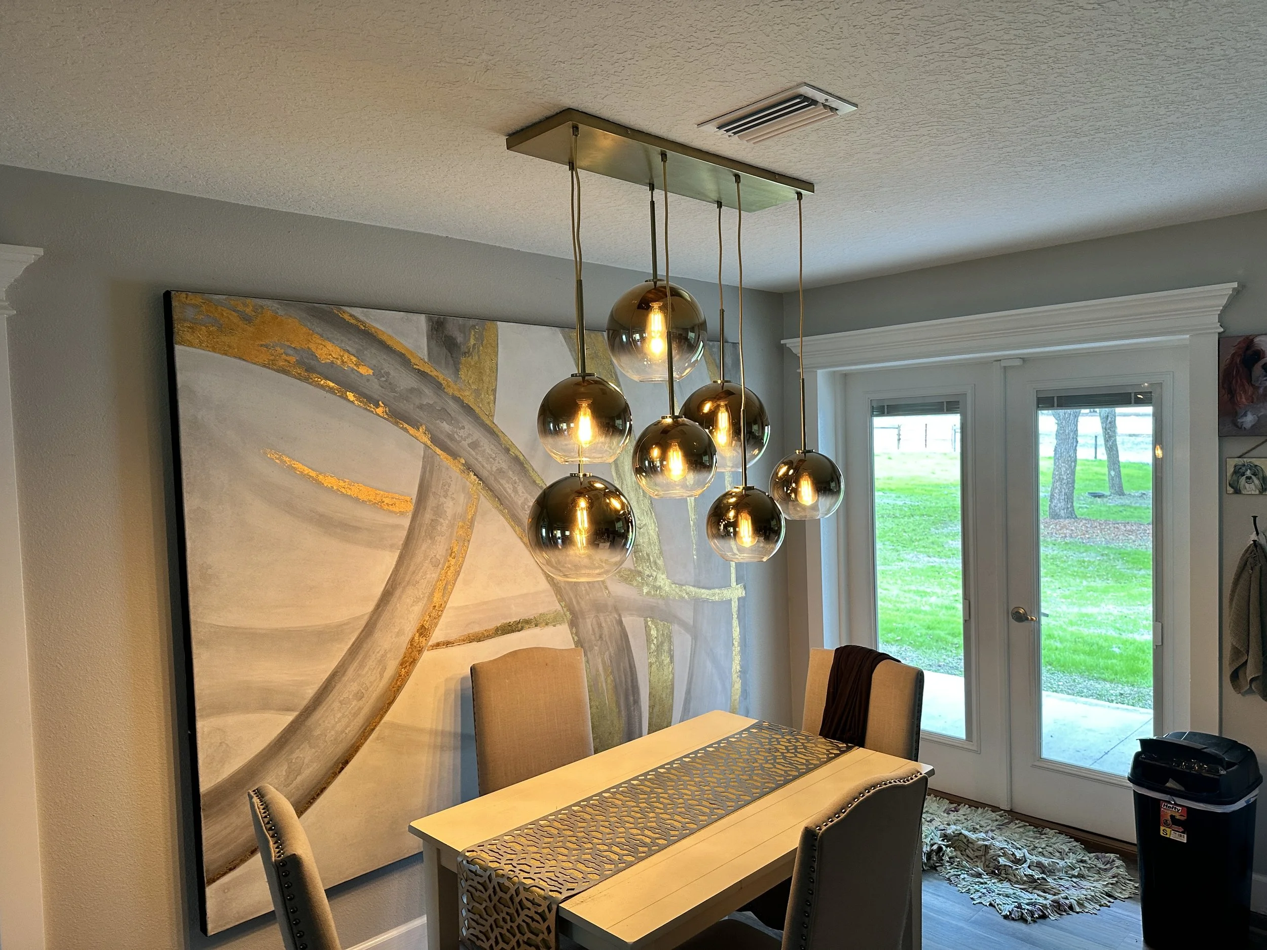 Modern dining room with a chandelier featuring multiple gold and glass globe lights, beige upholstered chairs, a large abstract painting on the wall, and glass double doors opening to a green backyard.