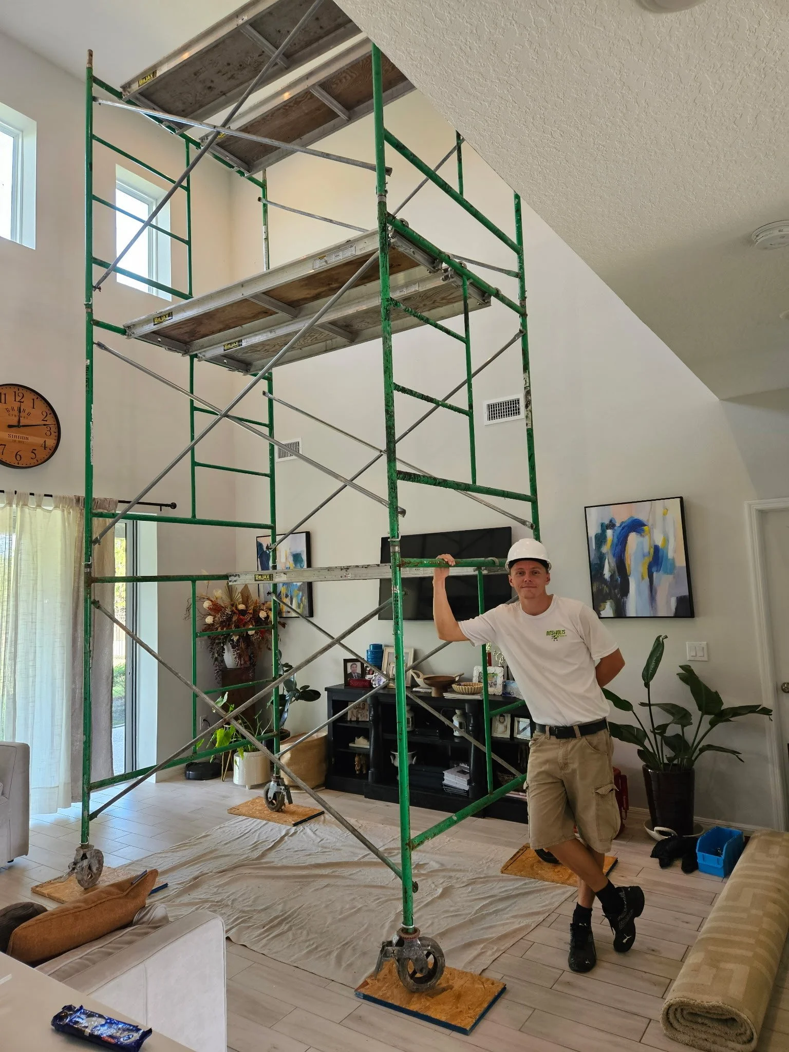 A man stands next to a large green and silver scaffolding inside a living room. The room has a beige rug, a black entertainment center, wall art, and large windows. The man is wearing a white helmet, white T-shirt, and tan shorts.