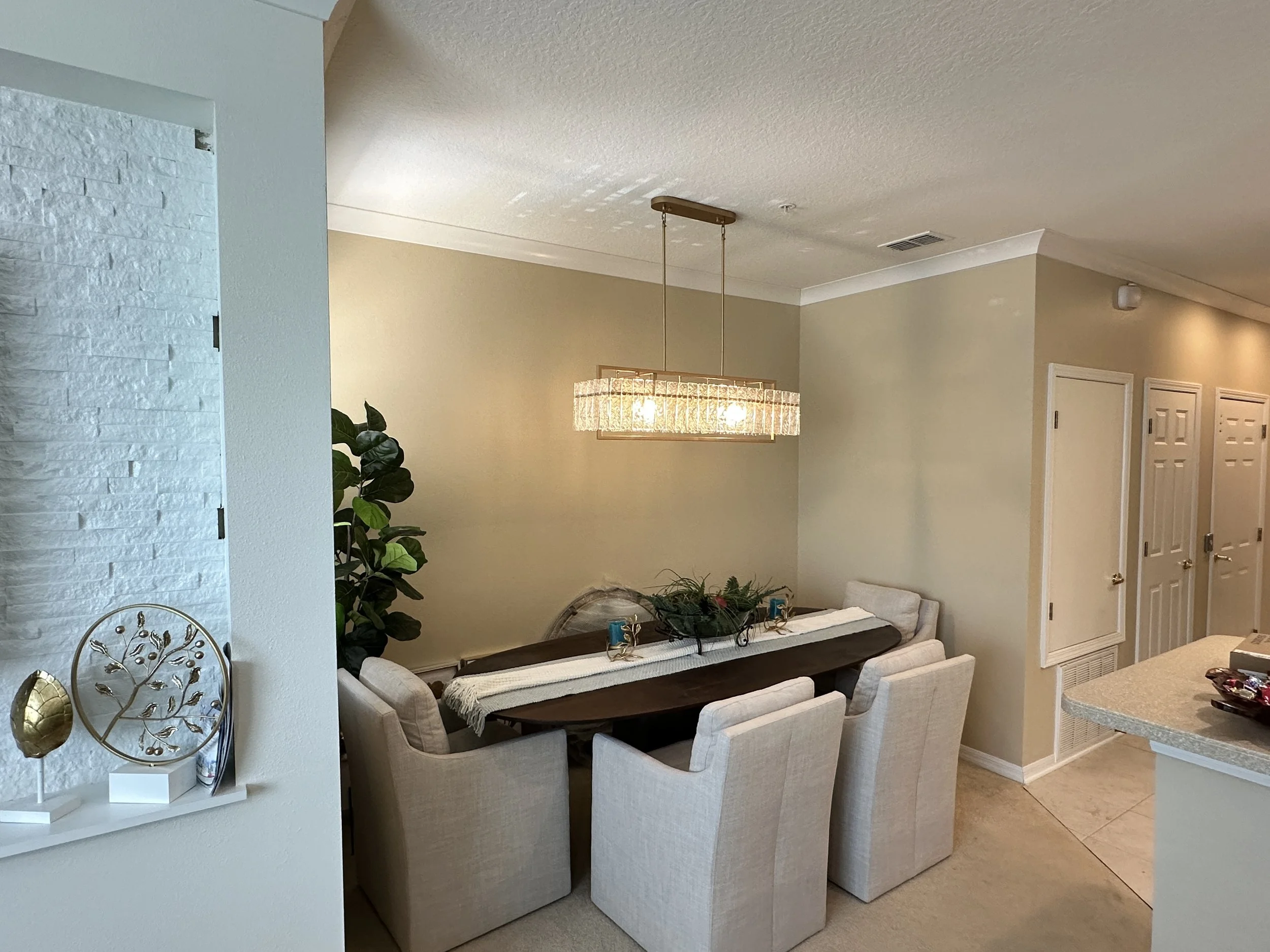 Dining room with a wooden table surrounded by six beige upholstered chairs, a modern crystal chandelier hanging above, a large green potted plant, and holiday decorations on the table.
