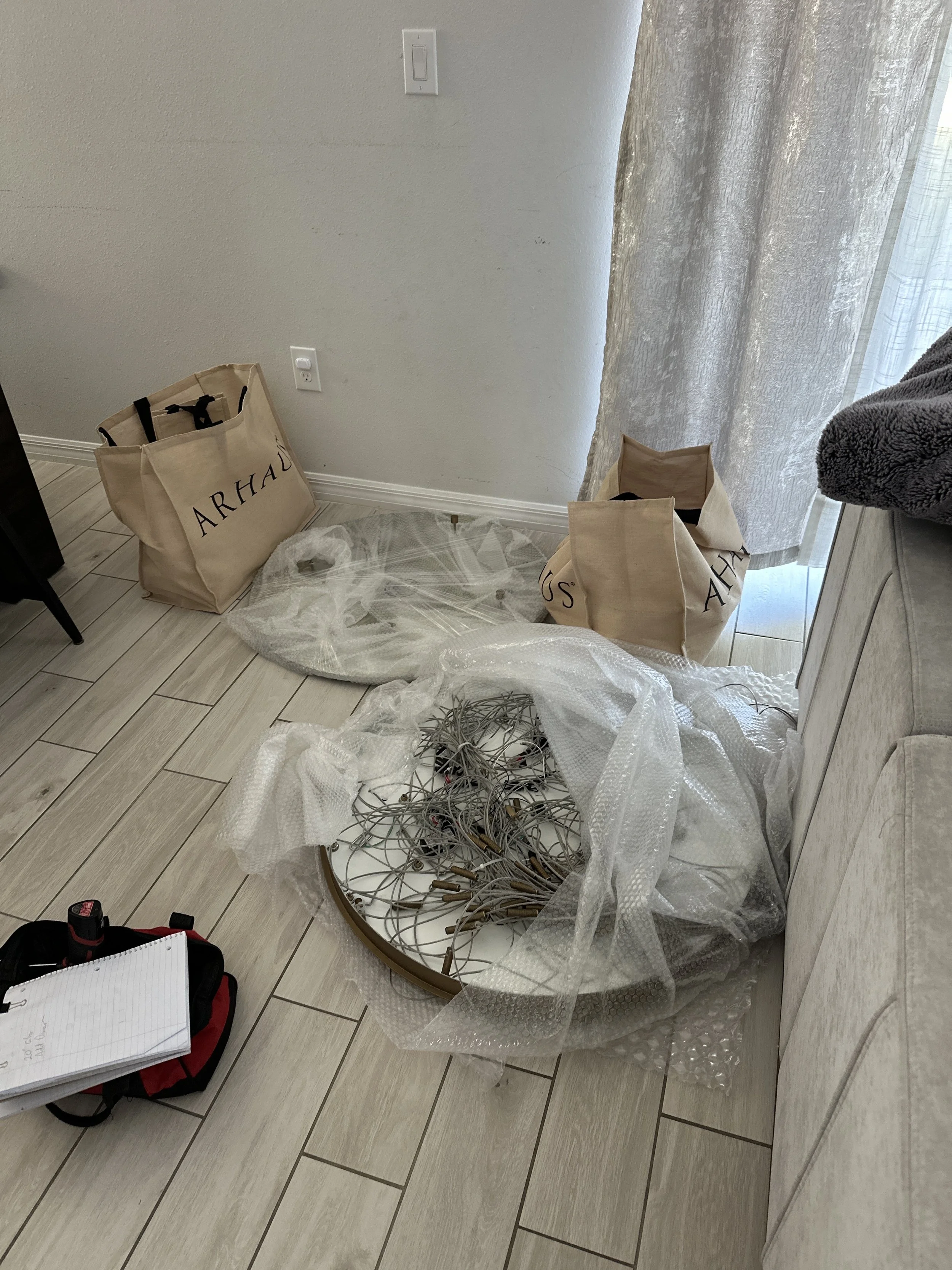 A corner of a room with two large shopping bags labeled "ARHAUS," a round table with tangled metal light fixtures wrapped in bubble wrap, and some paperwork with a flashlight on a black and red bag on the floor. There is also a remote control and notebook on top of the bag, beige tile flooring, and a gray curtain partially covering a window.