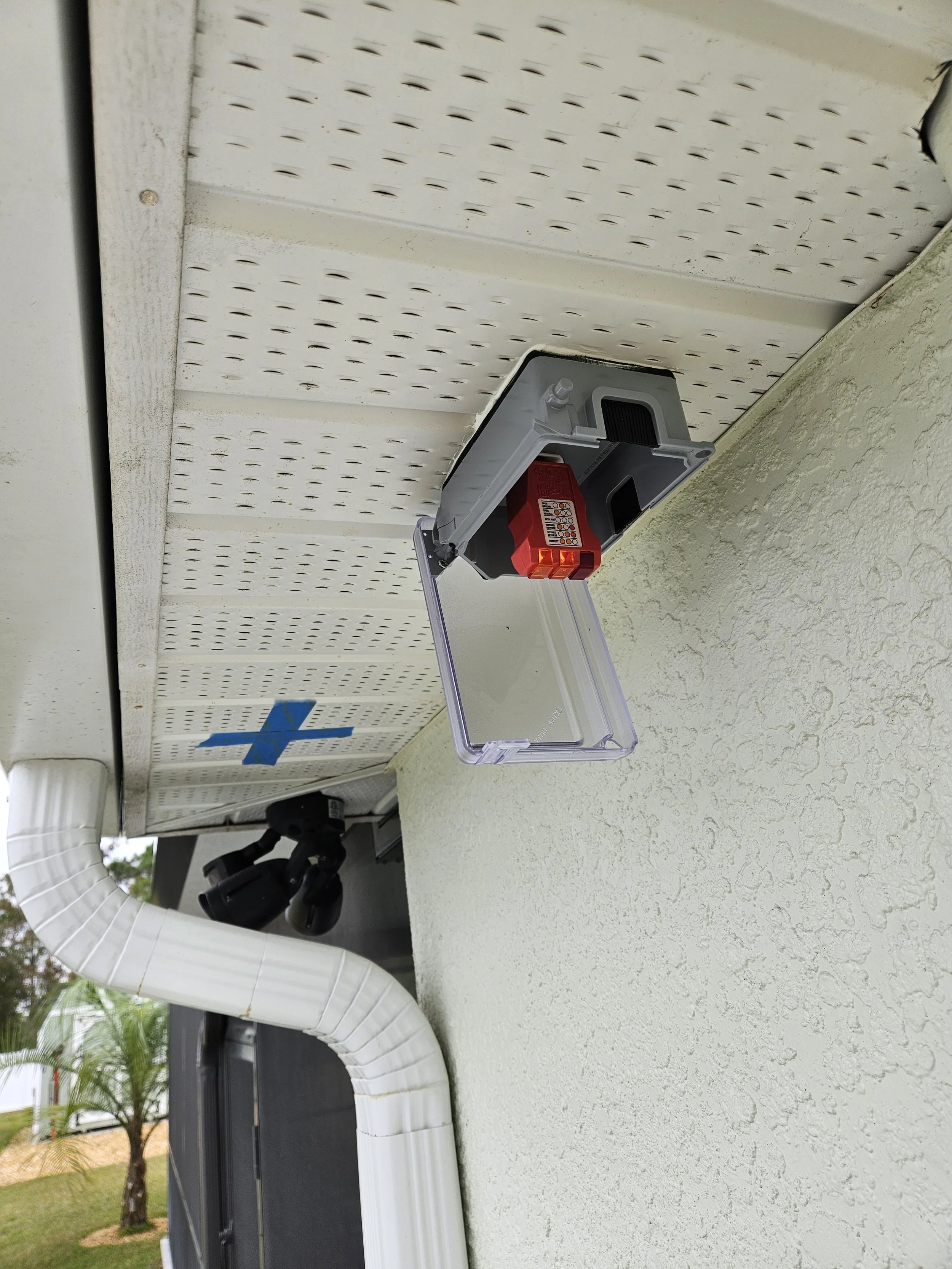 An outdoor wall with a partially open electrical or alarm box with a red component inside. Ductwork and a surveillance camera are also visible.