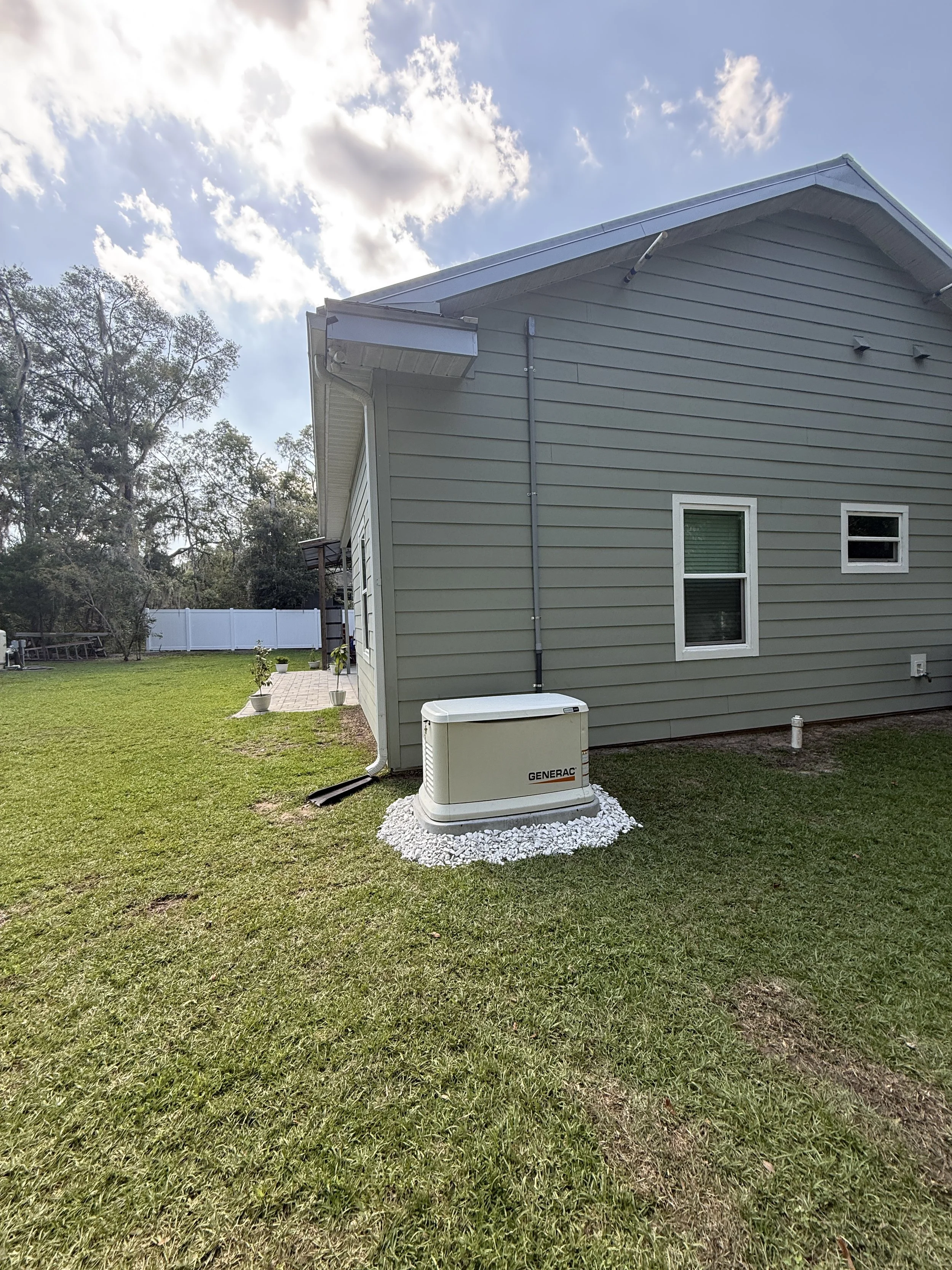 Backyard with green grass, house with gray siding, two small windows, a white fence, and an electrical generator on a bed of white rocks. Trees and cloudy sky in the background.