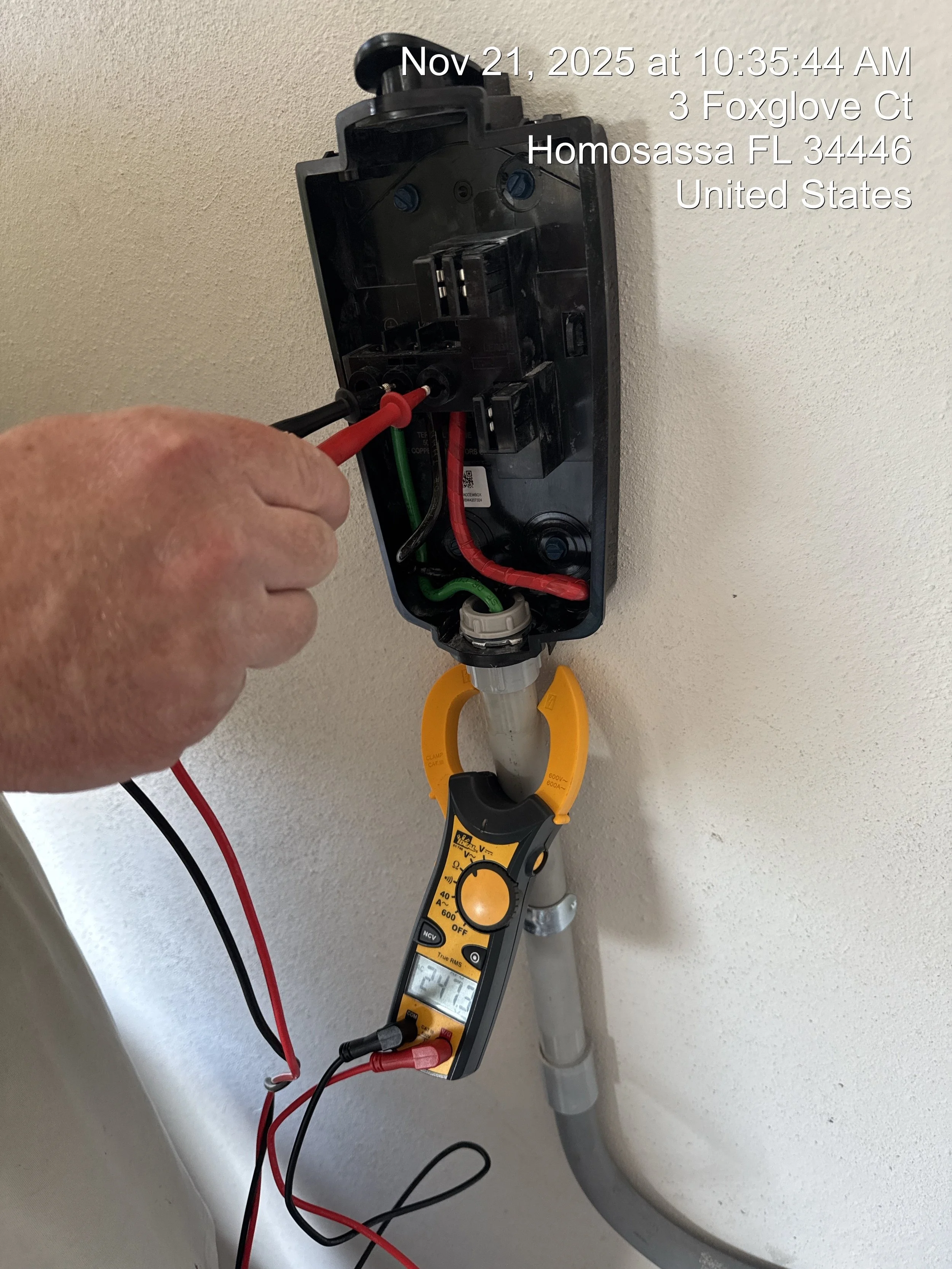 A person testing electrical wires in an electrical box using a digital multimeter. The setup includes red and black test leads connected to the wires and a multimeter displaying a reading, with a pipe below the box on a wall.
