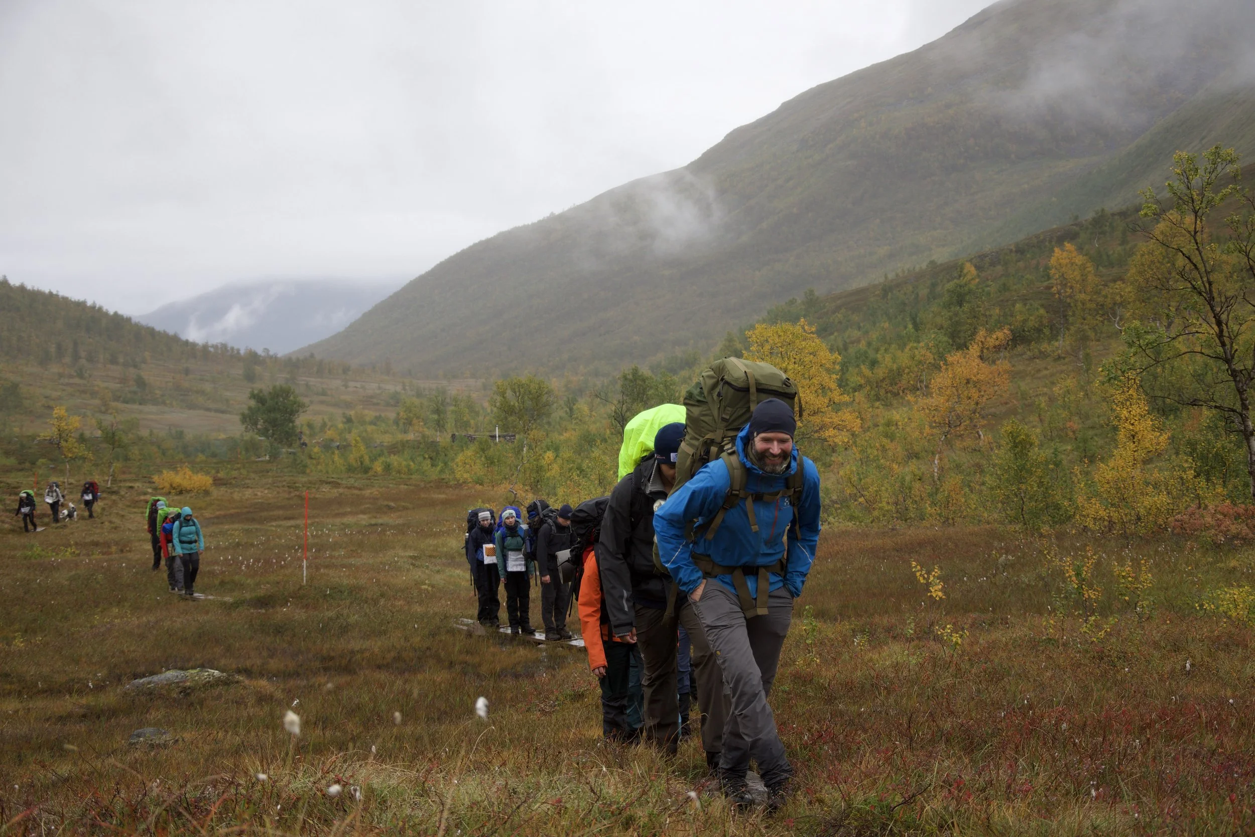 Grupp mennesker går gjennom et åpent landskap med grønne enger og steiner, omgitt av fjell og høyere fjelltopper med snø avsides, under en overskyet himmel.