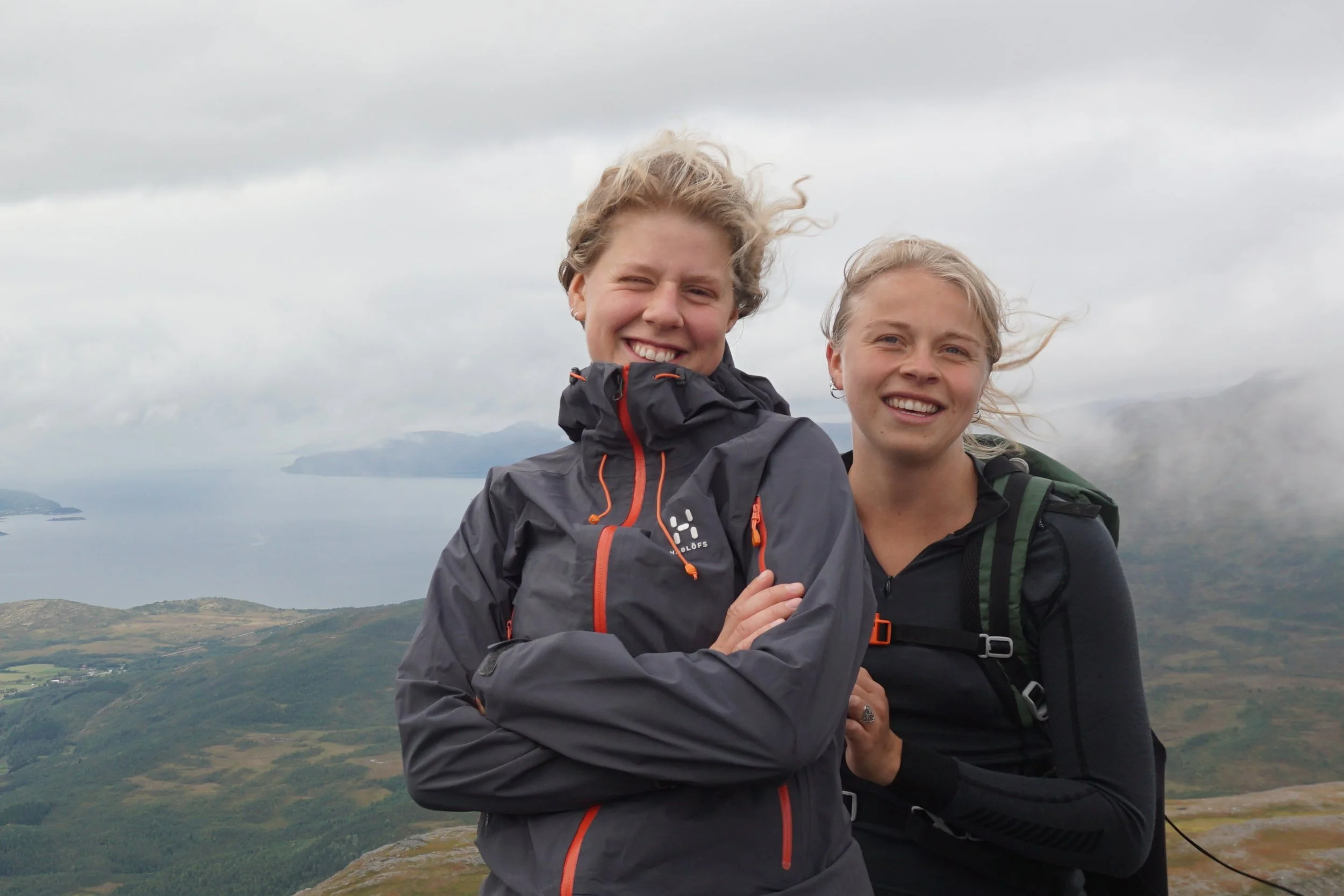 En gruppe unge mennesker med ryggsekker på ryggen står sammen i fjellandskap med vann og fjell i bakgrunnen. De har hjelmer og utstyr for turgåing, og noen av hundene ligger foran dem.