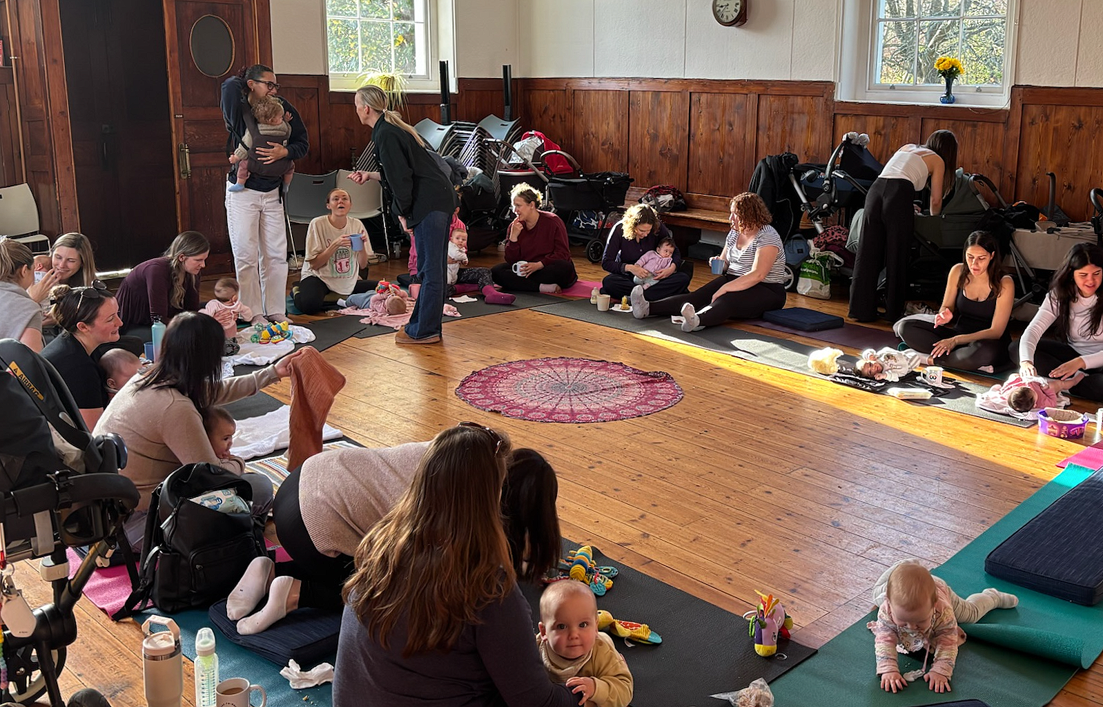 Children and adults at a gathering in a living room, some babies on the floor, some sitting on chairs and couches, with snacks and drinks on the table and walls decorated with artwork and shelves.