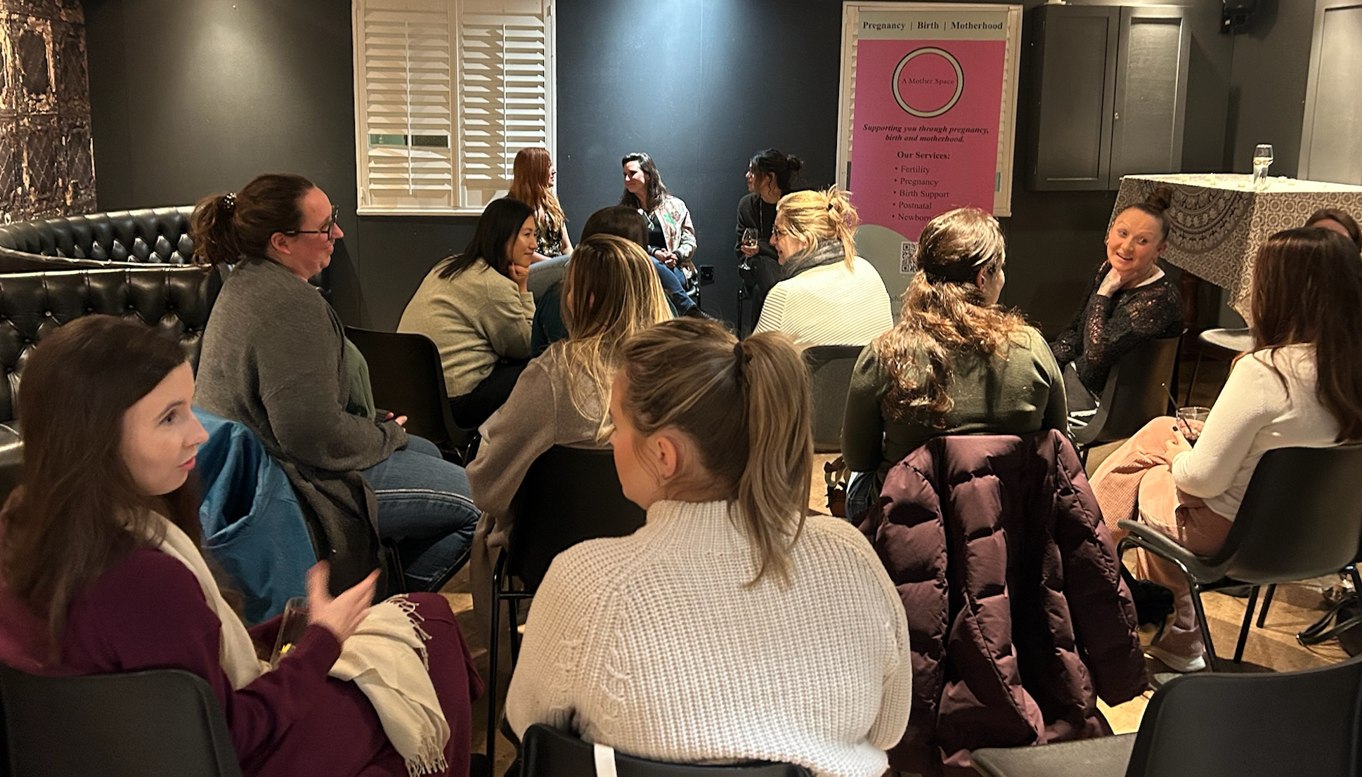 Group of women seated in a circle at a seminar or support group in a cozy room, some holding drinks, with a pink banner in the background about pregnancy and motherhood services.