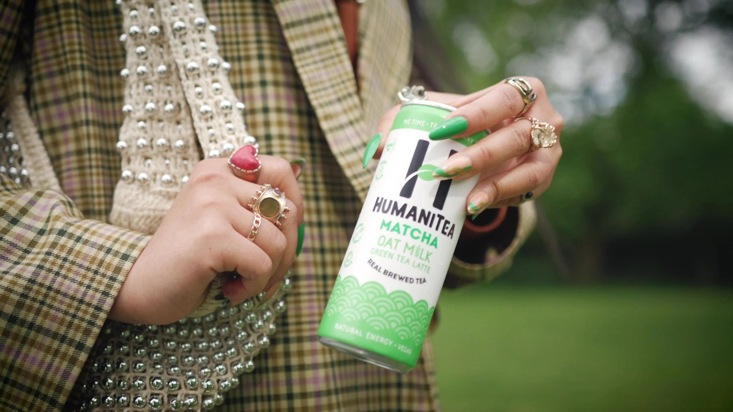 Person holding a can of Humanitea Matcha green tea latte outdoors, wearing multiple rings and a plaid jacket with pearl embellishments.