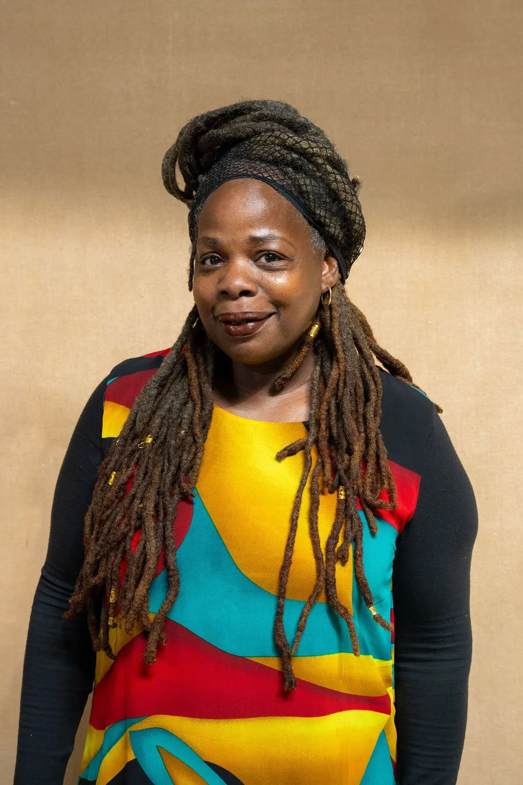 A woman with long dreadlocks wearing a colorful dress with a yellow, red, blue, and black pattern, and black sleeves, standing in front of a plain beige wall.