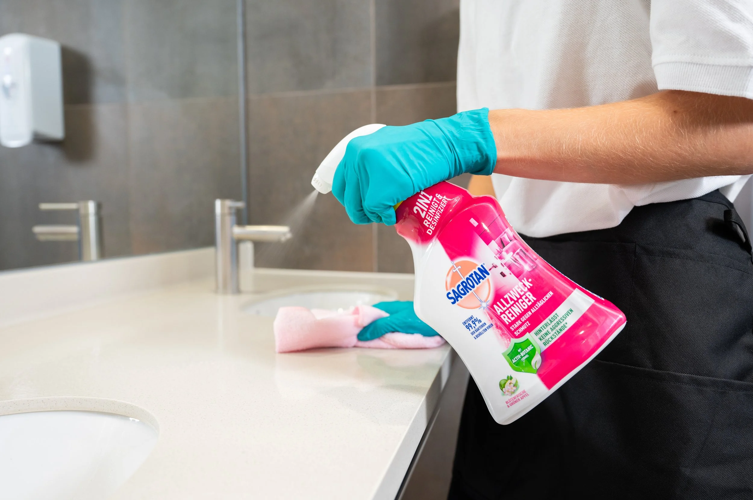 Person wearing blue gloves spray cleaning solution on a pink cloth at a bathroom sink.