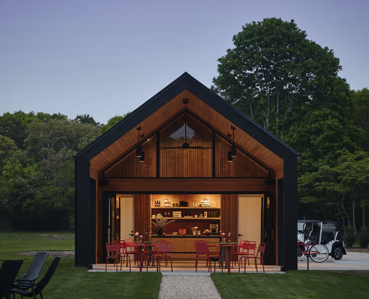 Modern open-concept wooden house with a high gabled ceiling, large glass doors, and a fully stocked kitchen, situated in a lush green outdoor setting during the evening.
