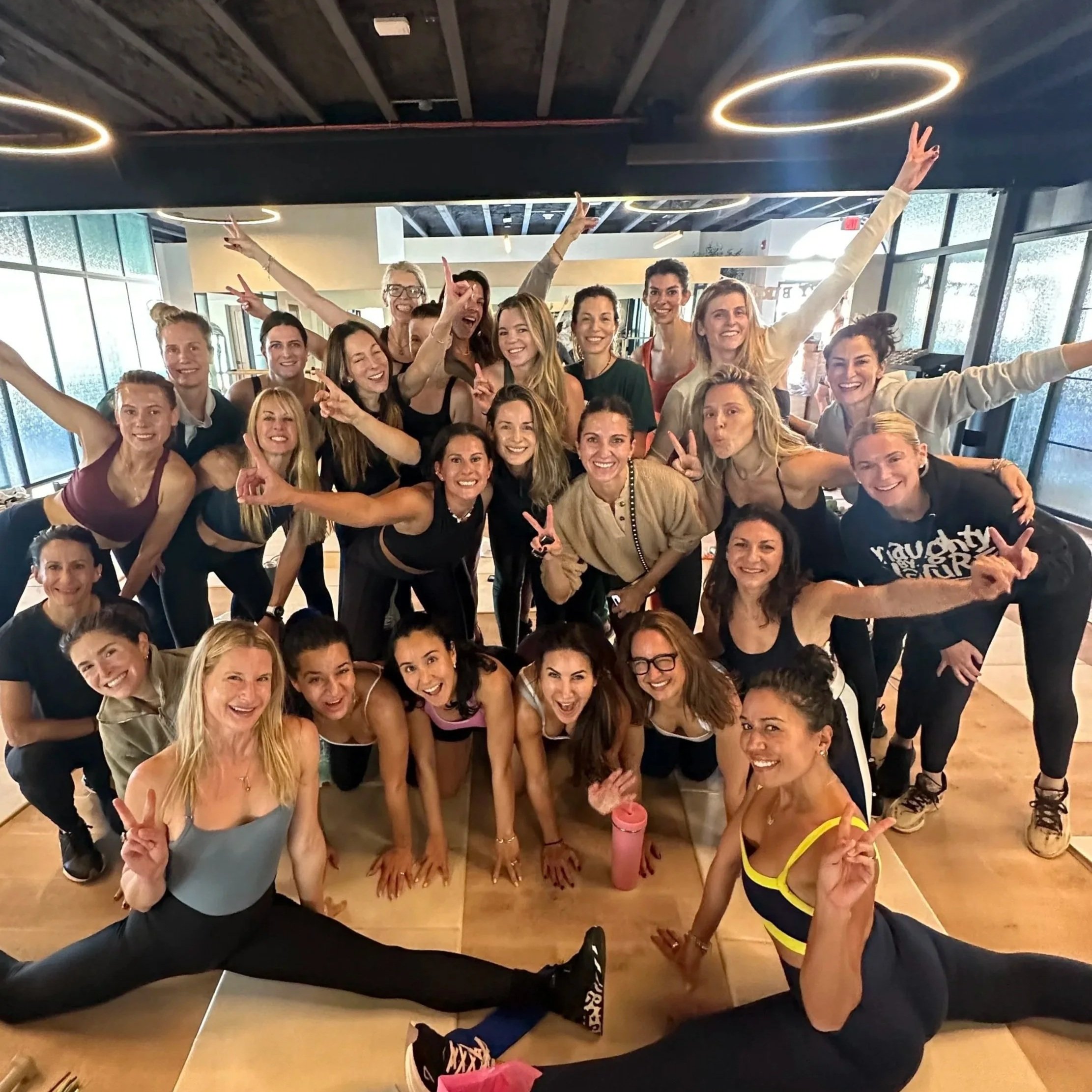 Group of women in workout attire in fitness studio, smiling, making peace signs, doing splits, posing for group photo.