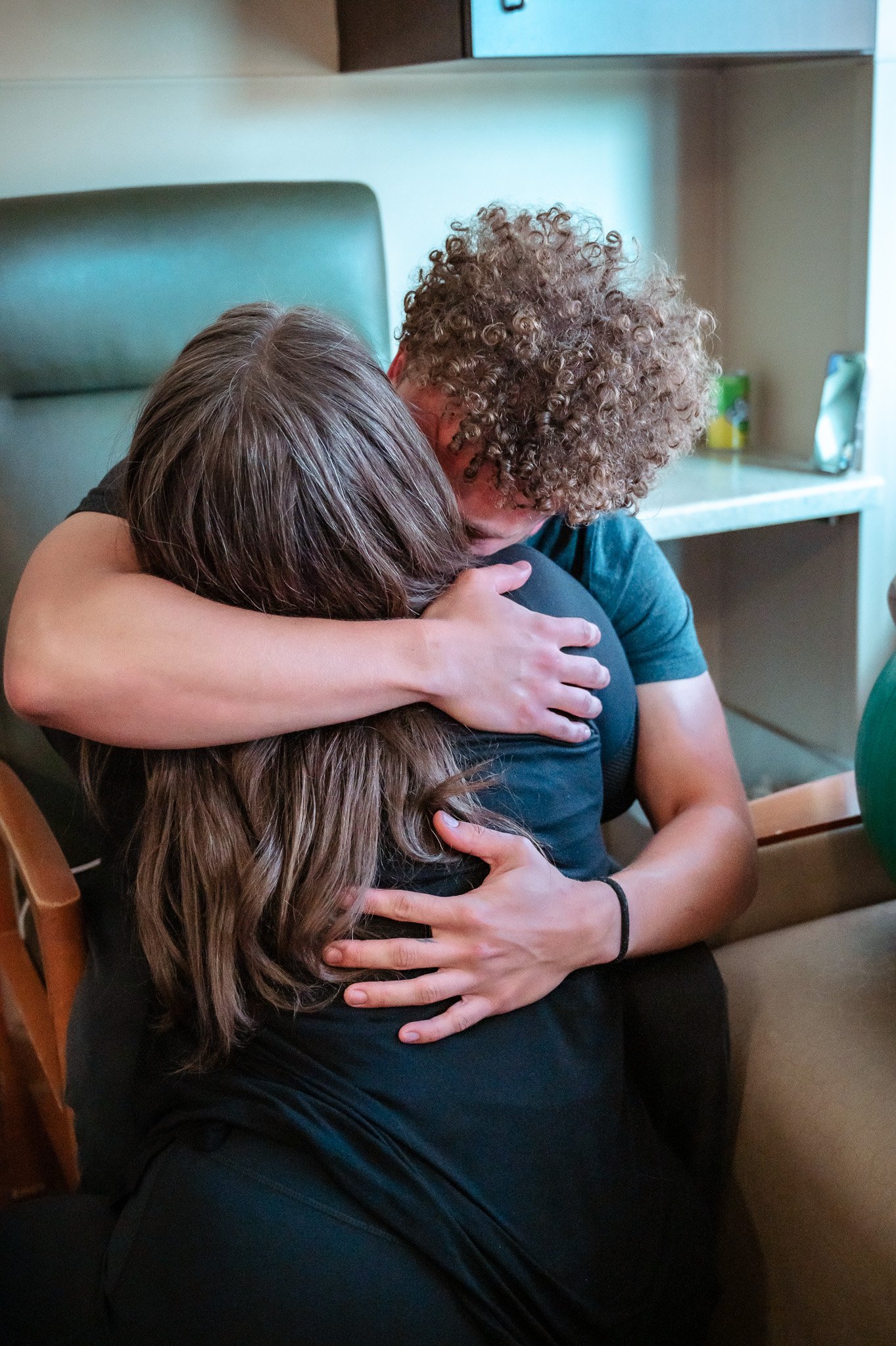 Two people hugging each other tightly in an emotional embrace, one with curly hair and the other with straight hair, in a hospital room.