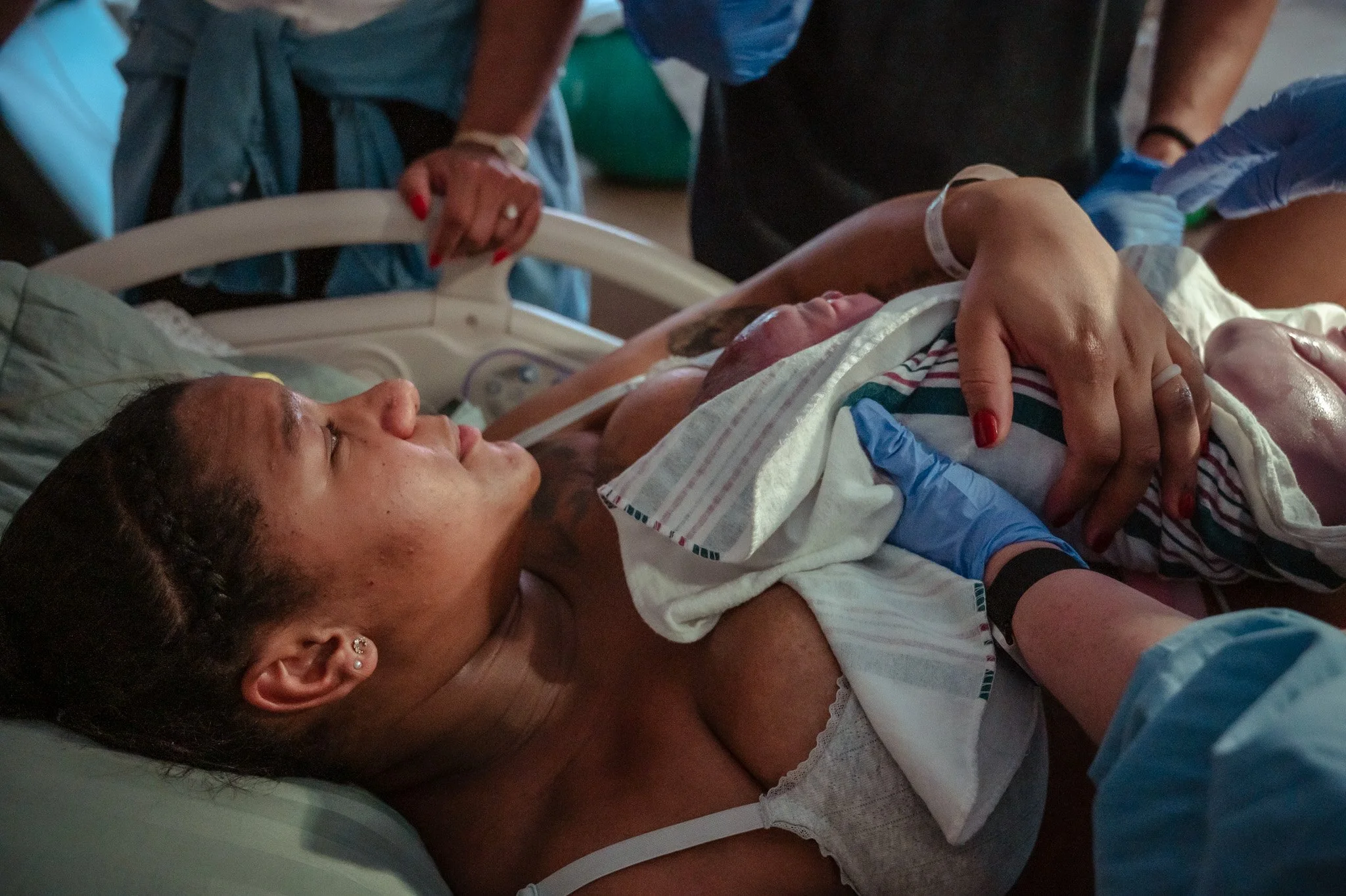 A woman lying in a hospital bed holding a newborn baby, with medical staff surrounding her. The woman appears emotional and the newborn is wrapped in a striped blanket.
