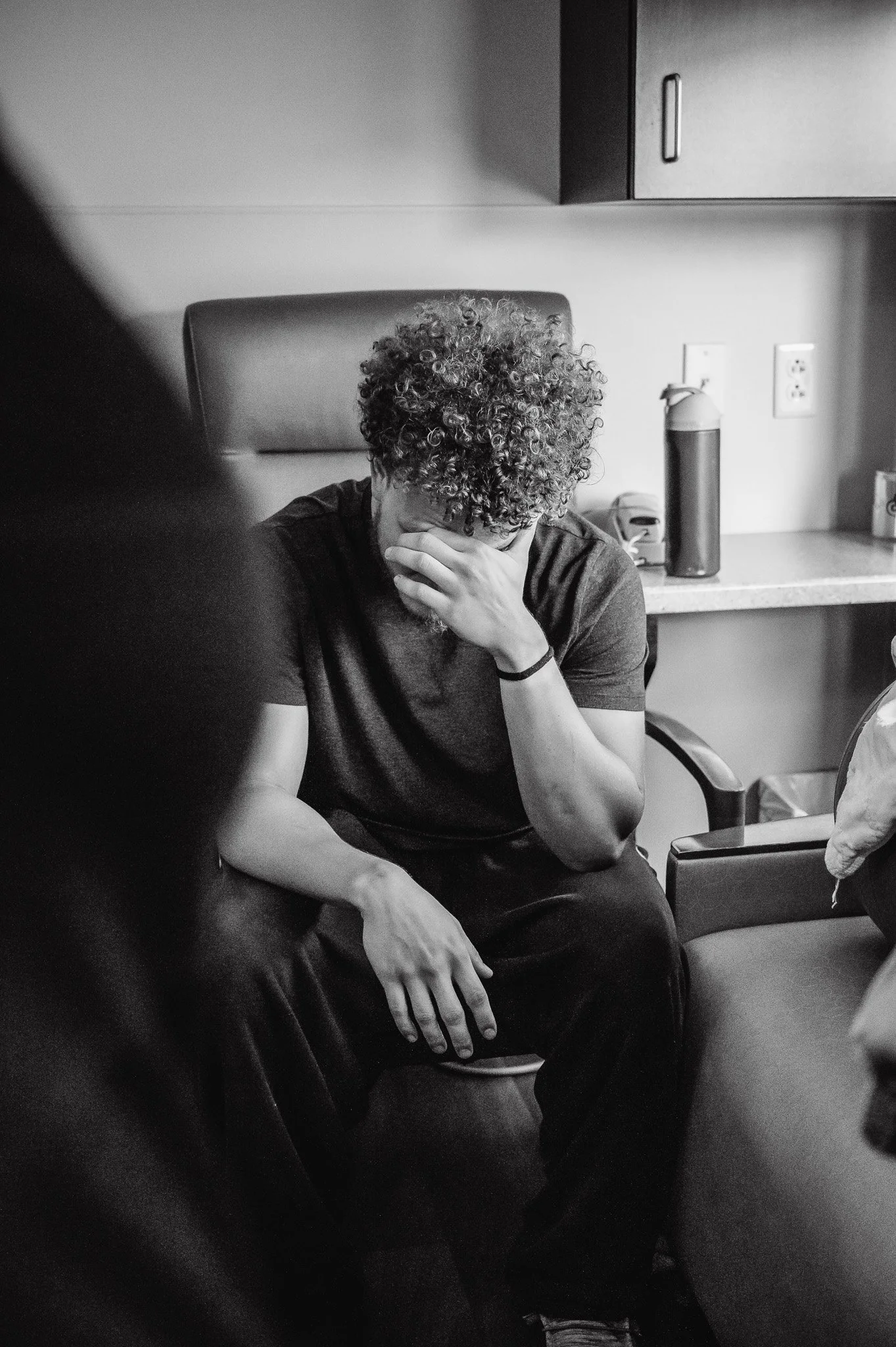 A person with curly hair sitting in a chair, covering their face with their hand, in a room with a countertop and cabinets in the background.