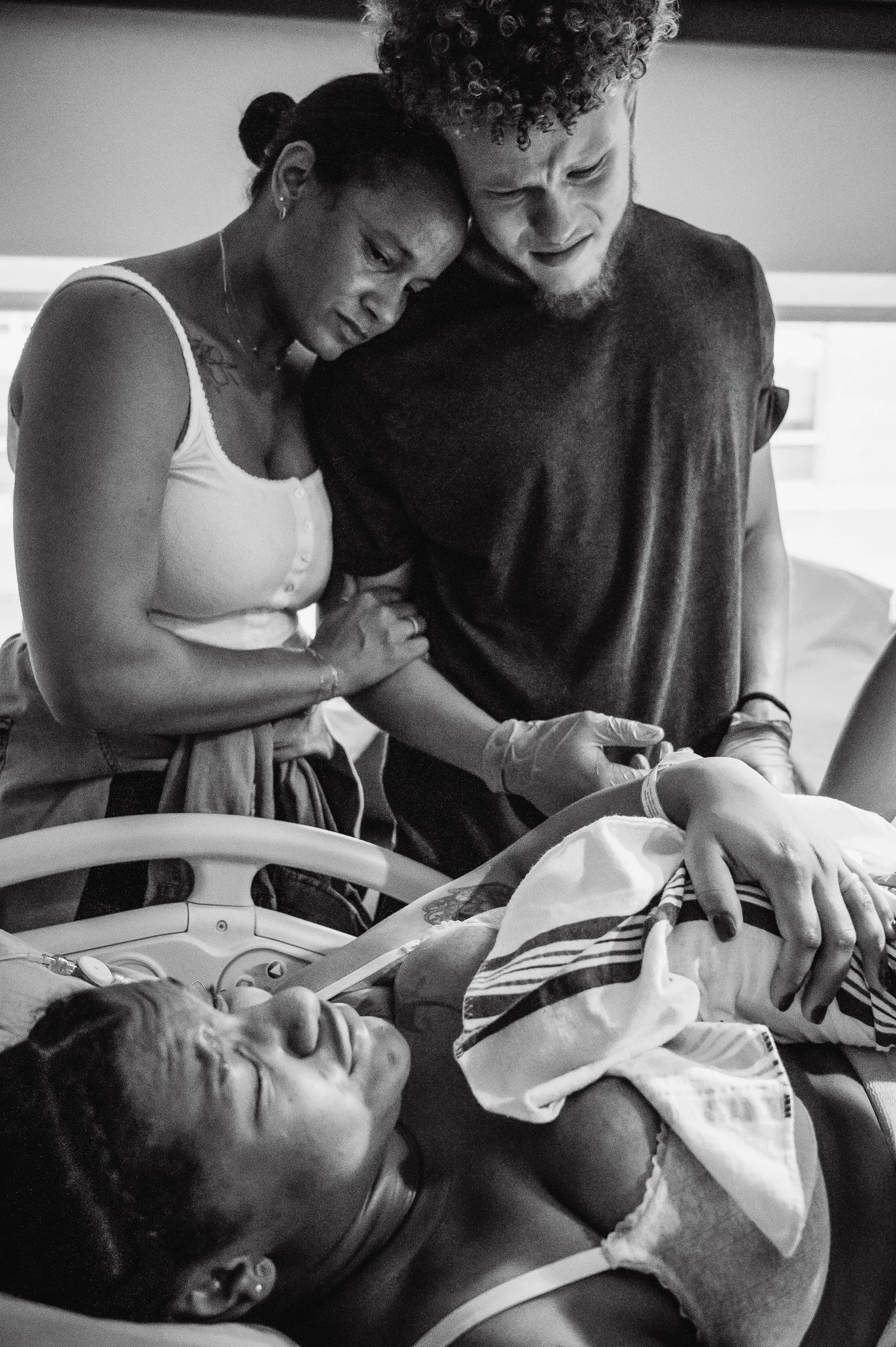 A woman and a man hold her arm as they look at a woman lying in a hospital bed, who appears to be in labor or has just given birth.