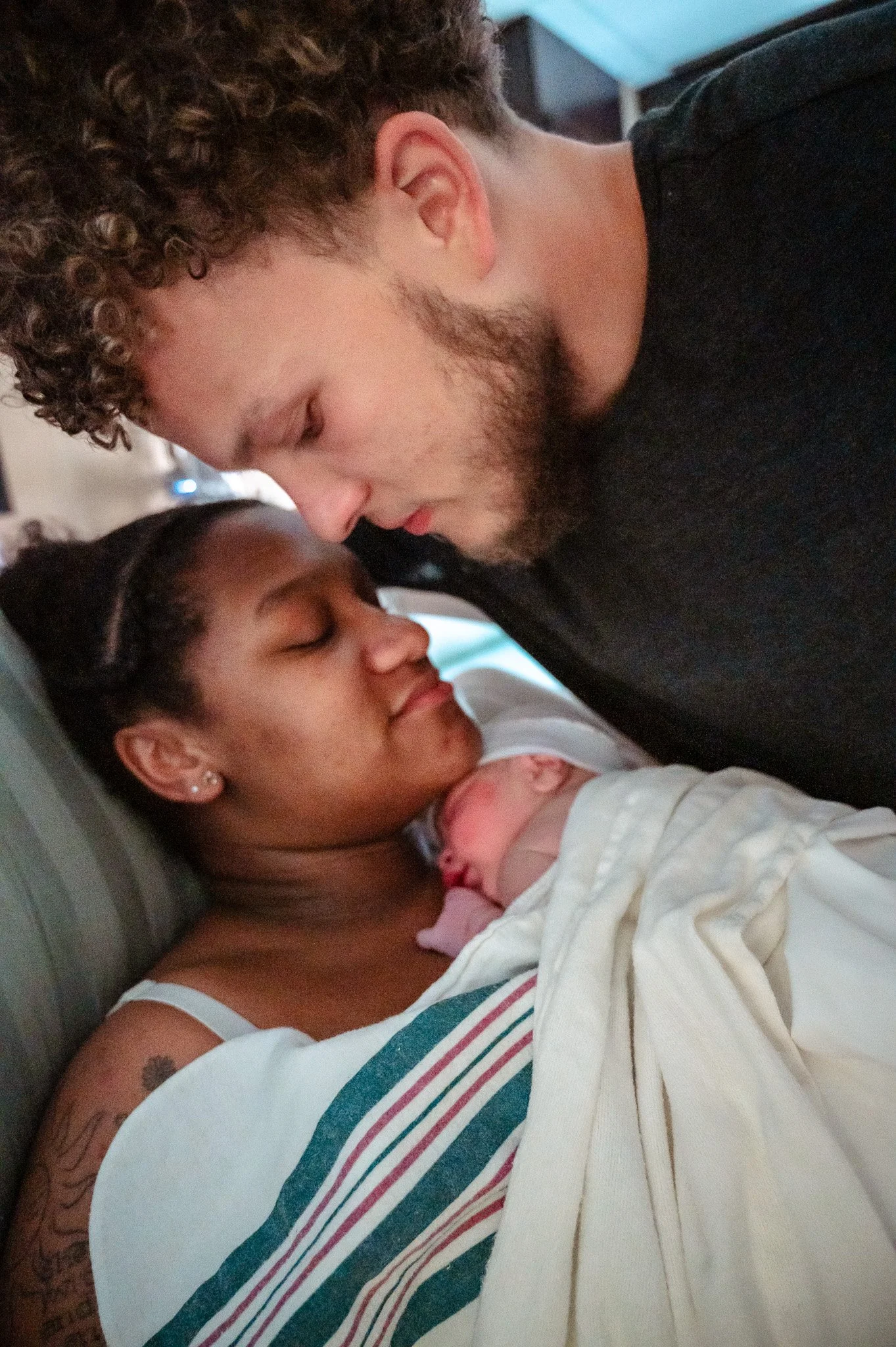 A woman lying in bed holding a newborn baby with a man leaning over her and touching foreheads in a hospital or maternity ward.