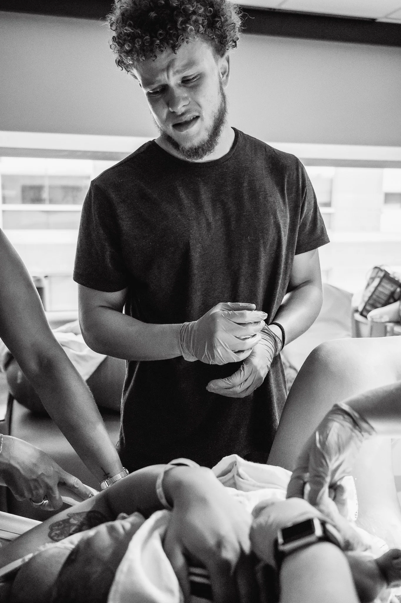 A man with curly hair and a beard, wearing a black T-shirt and gloves, is assisting a woman during childbirth in a hospital room.