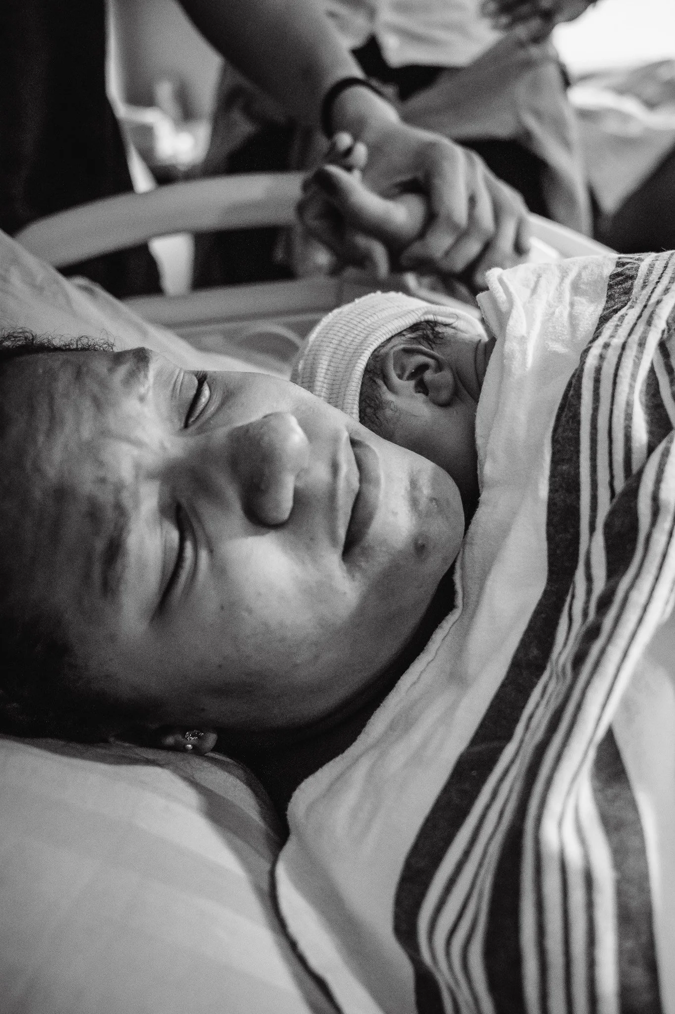 A woman lying in a hospital bed holding a newborn baby close to her chest. The woman appears to be resting with her eyes closed, and the baby is dressed in a striped outfit and hat, resting peacefully.