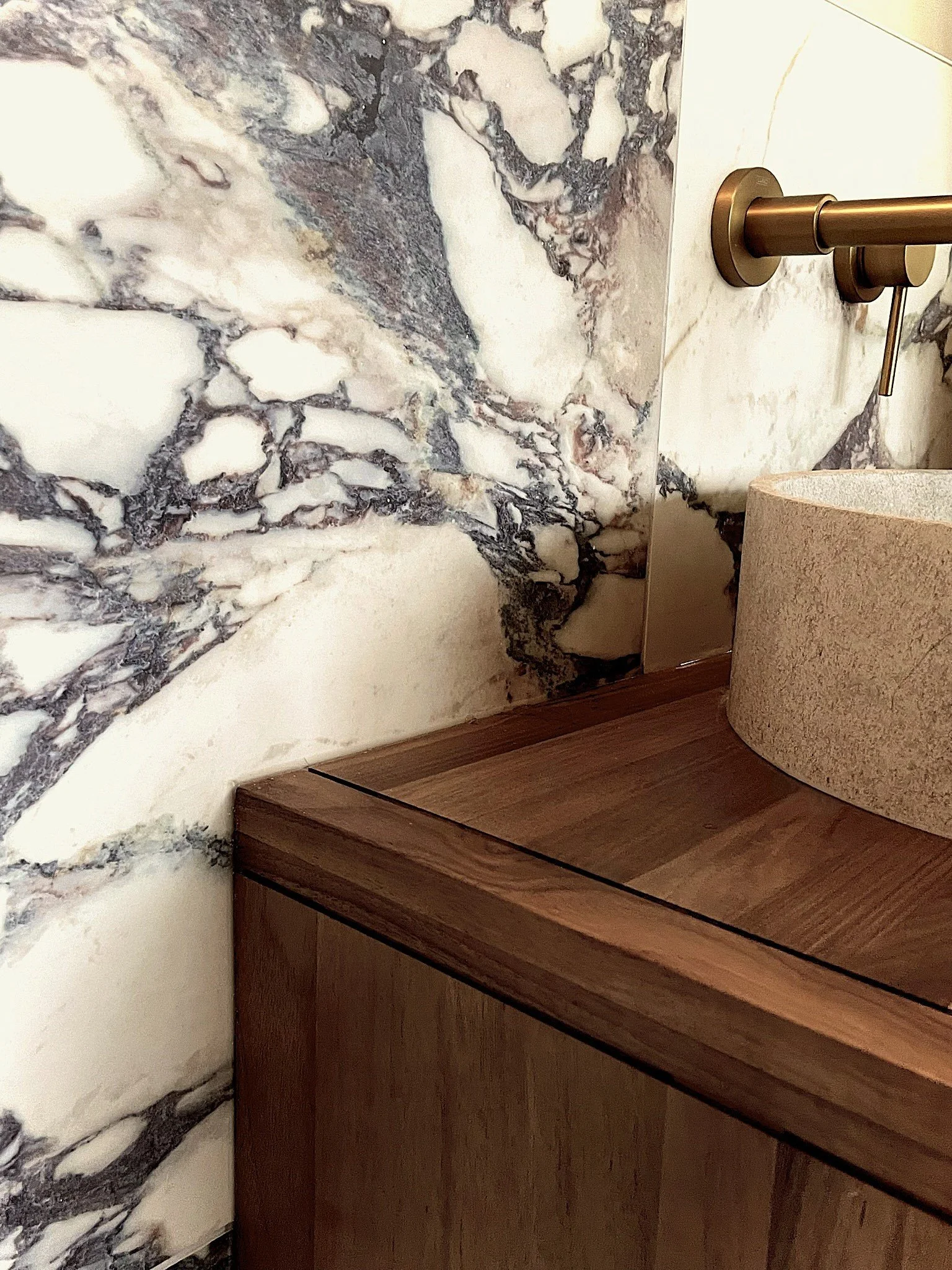 Close-up of a wooden bathroom vanity with a stone vessel sink, a marble wall with dark veins, and a metallic wall-mounted faucet.