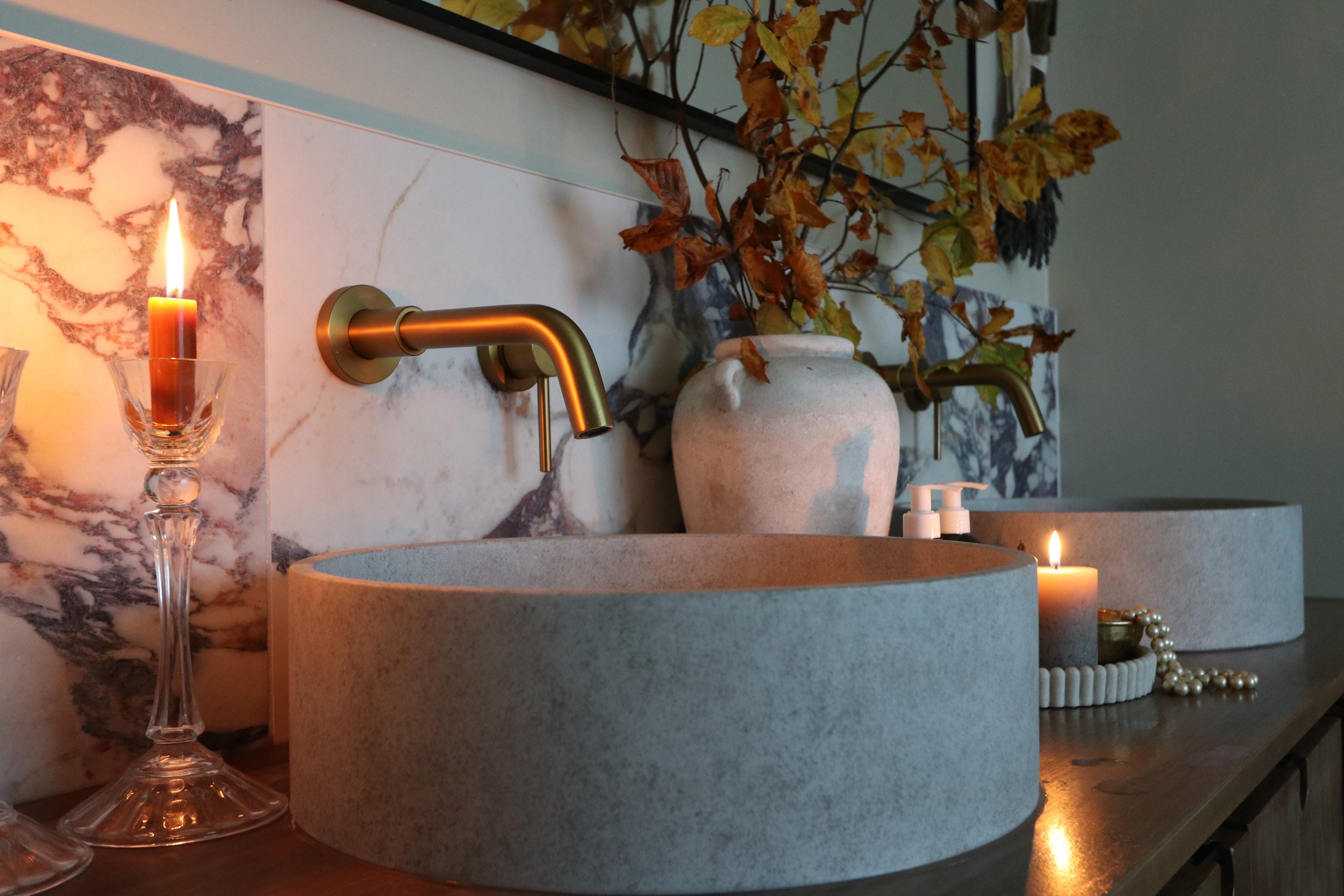 A bathroom countertop with two round stone sinks, two brass wall-mounted faucets, a marble backsplash, a beige vase with dried leaves, lit candles, a string of pearls, and a hand soap dispenser.