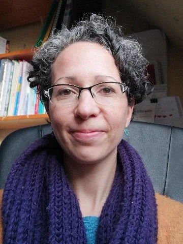 Une femme aux cheveux gris, portant des lunettes, un pull violet et un t-shirt bleu, assise dans un bureau avec des étagères de livres.