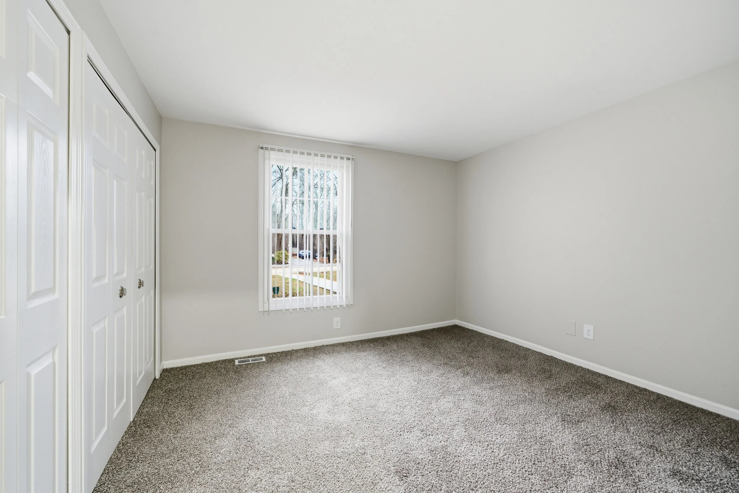 Empty bedroom with beige carpeting, white walls, a window with vertical blinds, and a closet with double white doors.