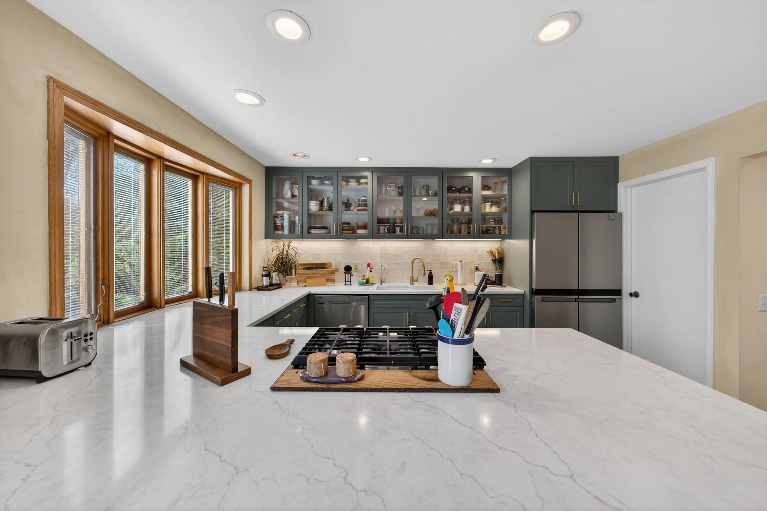 Modern kitchen with gray cabinets, white marble countertops, and a stainless steel refrigerator. Large windows with wooden trim let in natural light. Items like toaster, cutting board, and utensils are on the counter.