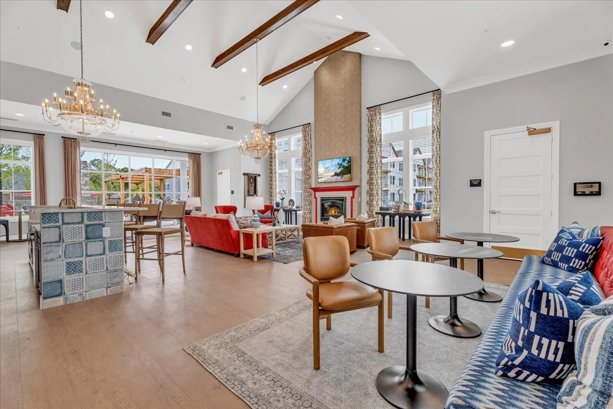 Bright and spacious living room with high sloped ceiling, exposed wooden beams, two chandeliers, large windows with floral curtains, a cozy fireplace with a TV above, various seating including red armchairs, a blue patterned sofa with cushions, a woo