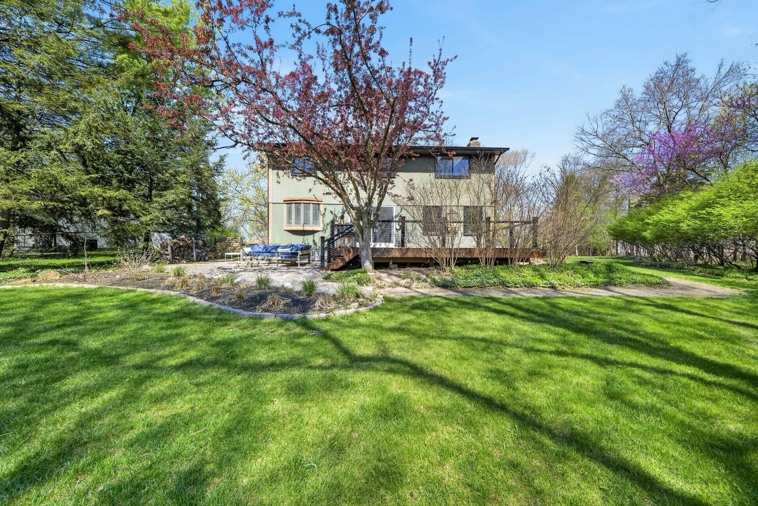 A backyard with a green lawn, colorful trees including pink and purple blossoms, and a two-story house in the background. There is a deck with outdoor furniture on the house.