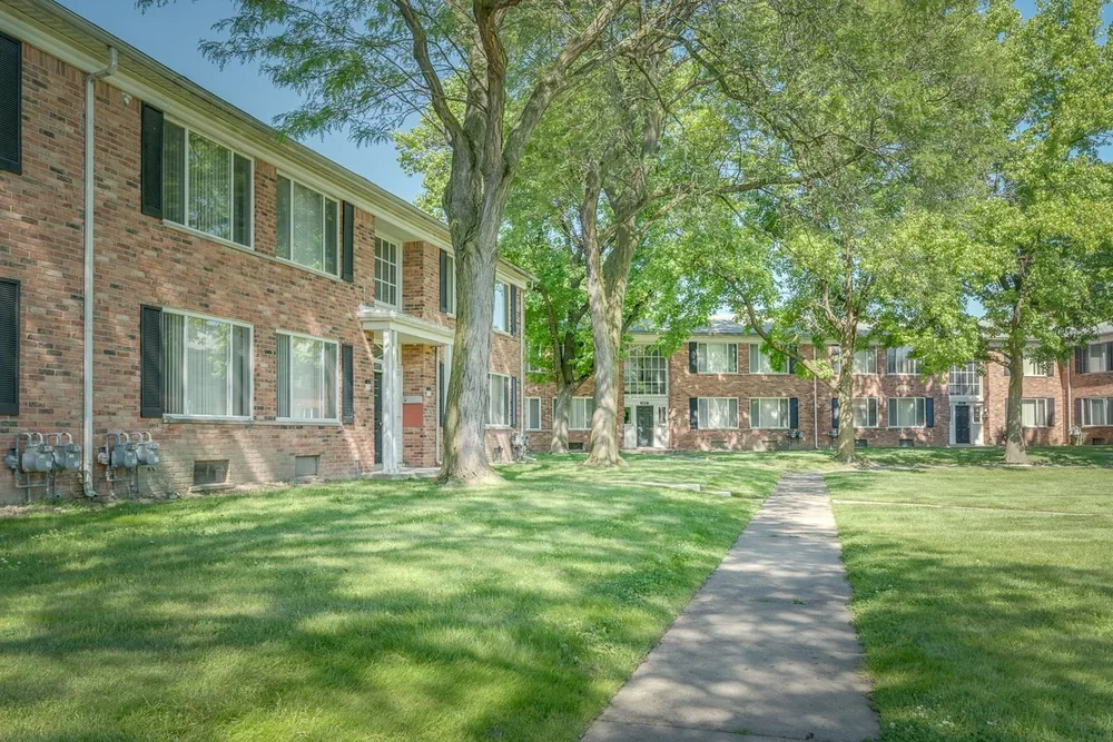 A brick apartment complex with multiple windows surrounded by green grass, trees, and a concrete sidewalk.